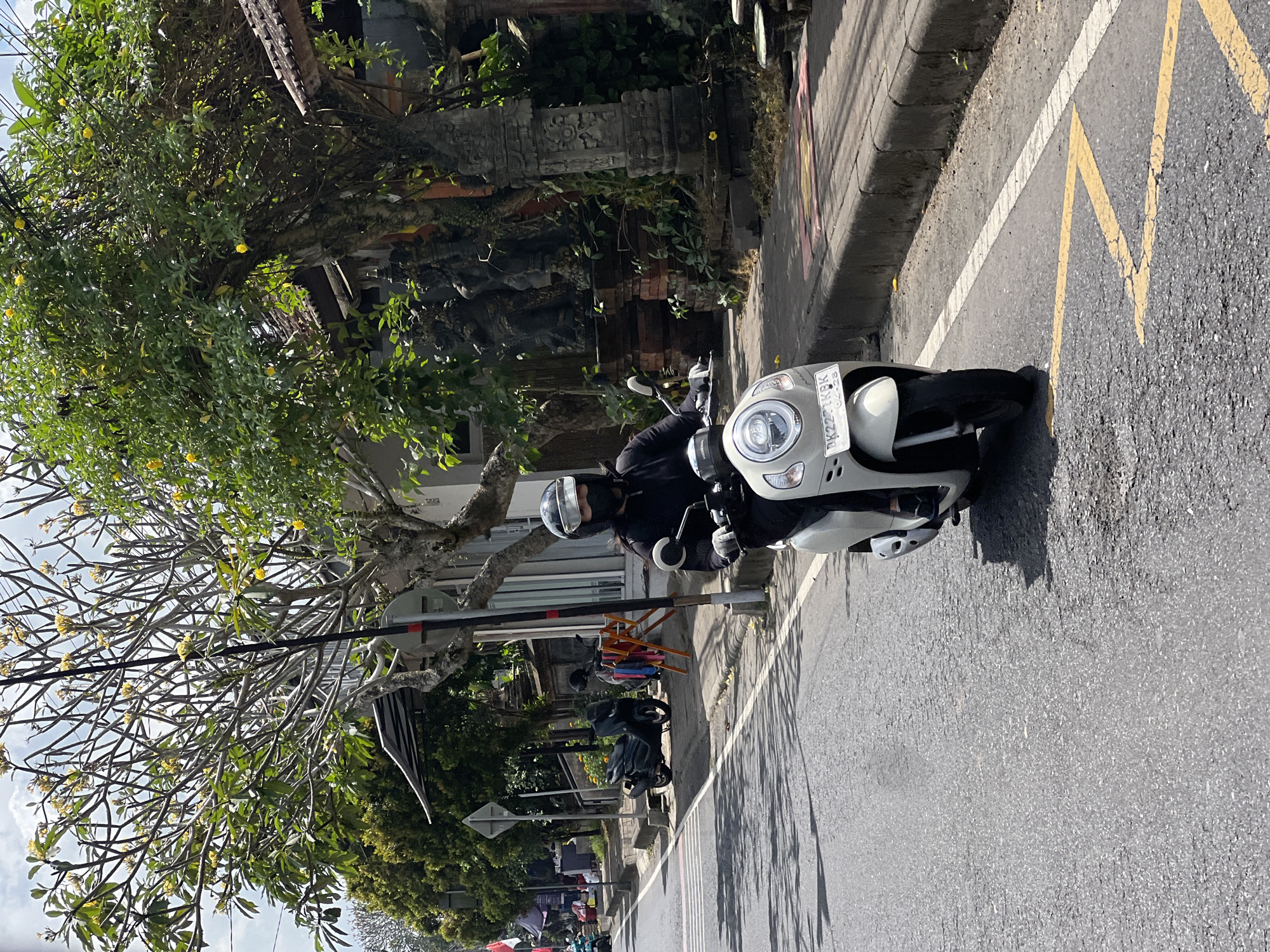 An individual wearing a black jacket, black mask, and a helmet with a clear visor rides a white scooter down a paved road in Payangan, Indonesia. The scooter has a license plate that reads "DK 2271 KBK". Yellow lines are painted on the road, and a white dashed line runs parallel to the curb. To the right of the road, there is a sidewalk with a curb, and beyond that, a property with a tiled roof, trees with yellow flowers, and stone statues. In the background, on the left side of the road, there are more trees and a few parked motorcycles. The sky is partly cloudy.