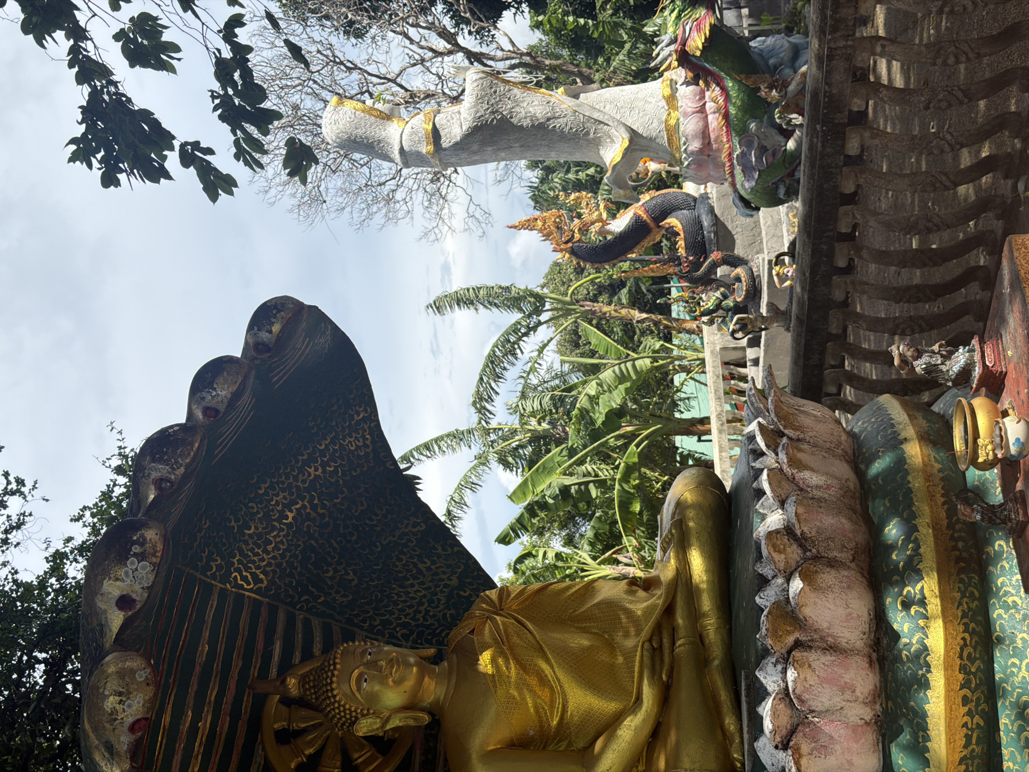 The image shows a collection of religious statues in an outdoor setting, likely a temple or shrine. In the foreground on the left, a golden Buddha statue is seated on a lotus flower base. Behind and to the right of the Buddha, a tall, pale statue of a robed figure stands with hands clasped. In the mid-ground, there are several mythical creature statues, including a black Naga (serpent) with a golden headdress, and a green dragon-like creature. The background features lush green vegetation, including banana trees, and a cloudy sky. The overall scene appears to be in a tropical environment. This scene is located at the Big Buddha Temple, also known as Wat Phra Yai, on Koh Samui island, Thailand, not in Rawai, Thailand.