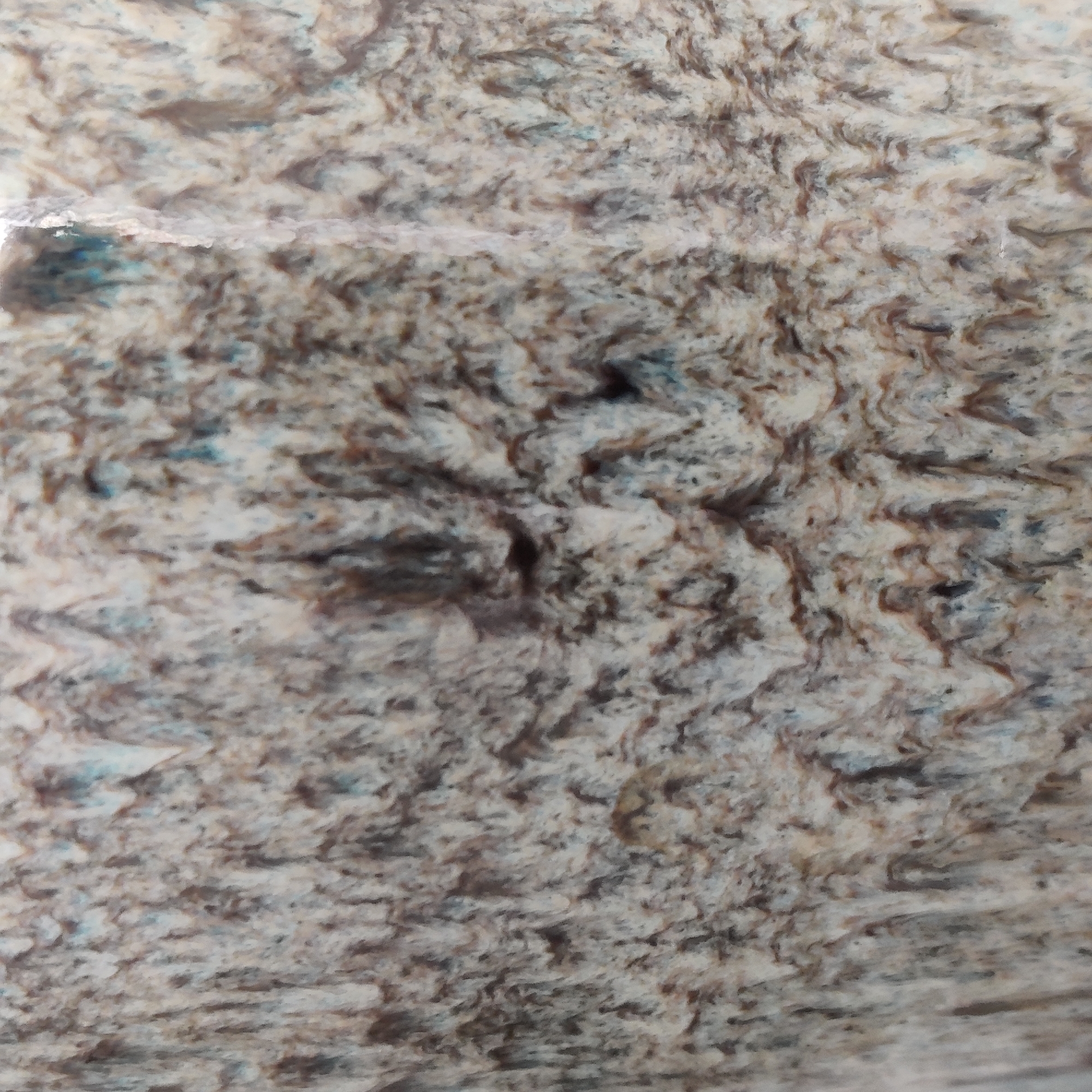 The image displays a close-up, abstract texture with a rough, variegated surface. It is predominantly composed of swirling patterns of light brown, grey, and white, with occasional hints of light blue. The texture appears to be reminiscent of natural stone, such as granite or marble, or perhaps a distressed ceramic material. The lighting creates subtle shadows that emphasize the raised and recessed areas of the surface, giving it a three-dimensional quality. There are no discernible objects, people, or environmental elements that would allow for identification of a specific location within Yekaterinburg, Russia, or any other city. The image is purely a study of texture and pattern.