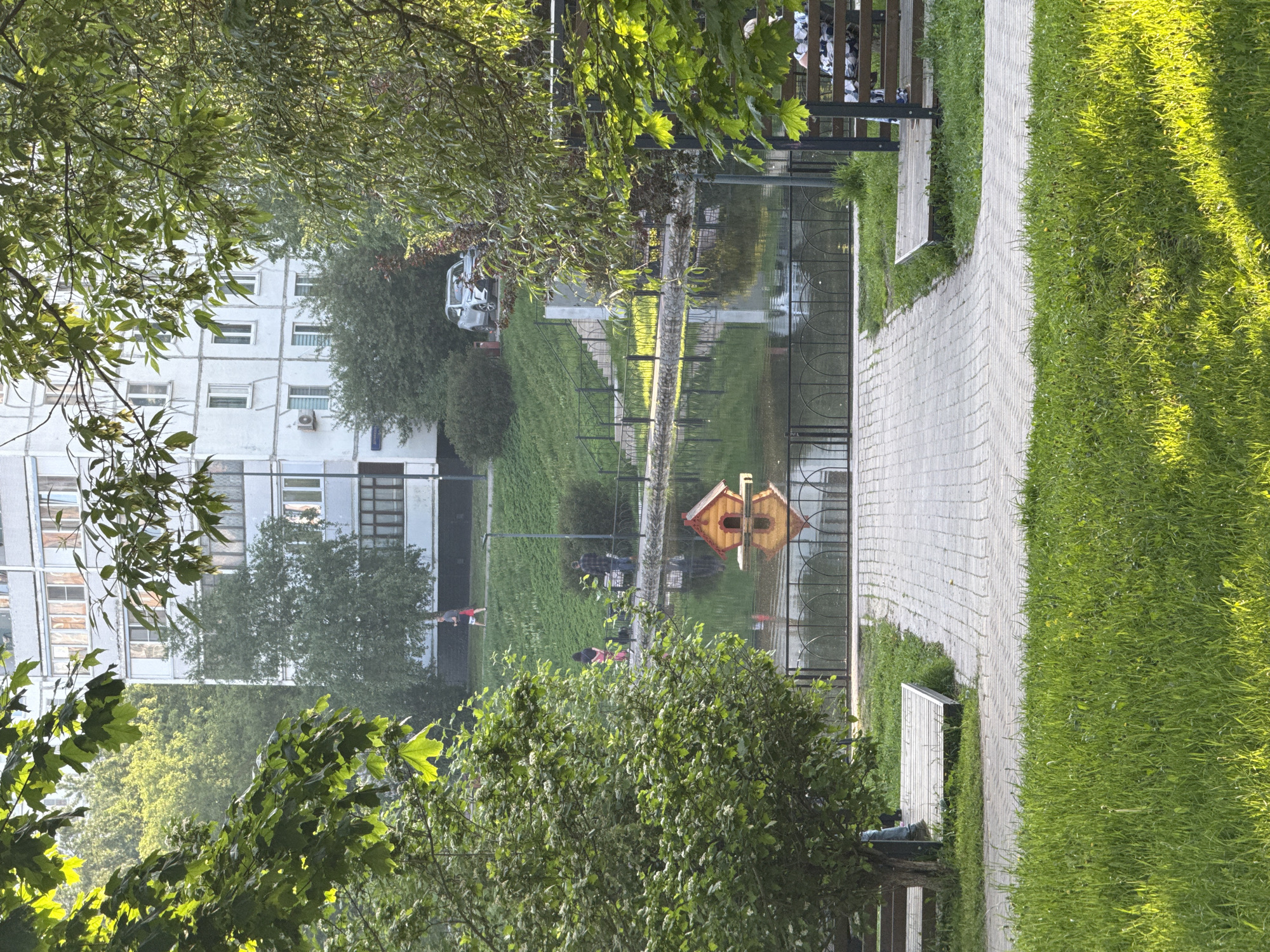 The image depicts a park in Moscow, Russia, with a pond in the center. A wooden birdhouse is situated in the pond. A paved walkway runs alongside the pond, with grass and benches on the right side. People can be seen walking in the background near a building, and a car is parked nearby. Trees are present throughout the park, framing the view. The sky is not visible.
