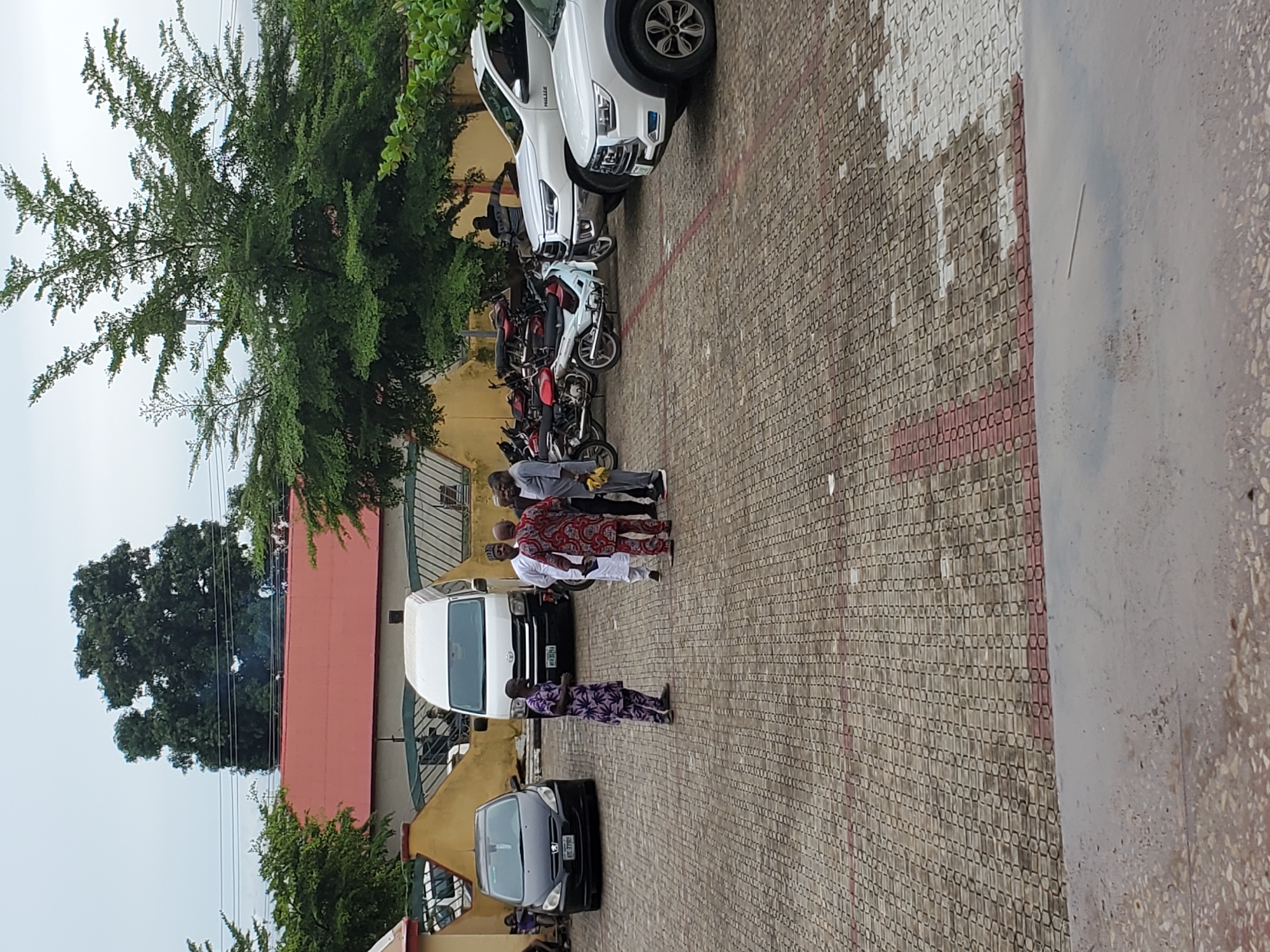 The scene depicts an outdoor paved area, likely a parking lot or courtyard, under an overcast sky in Mararaba, Nigeria. The ground is covered with interlocking concrete blocks patterned in light gray/beige with reddish-brown outlines, showing several wet patches or puddles reflecting the sky, particularly in the foreground.
Approximately seven to nine adult males are present. In the center-left, three men are engaged in conversation: one in a purple and white patterned traditional outfit (facing left), one in a white traditional outfit (facing the group), and one in a red and white patterned traditional outfit (facing the group). To the far right, a man in a light gray traditional outfit stands, holding a yellow object. Near a cluster of motorcycles, another man in a dark striped shirt is visible. Indistinct figures are also present near the buildings in the background.
Vehicles include a black Peugeot 307 sedan (license plate: KRC 770BB) parked on the far left, and a white Toyota Hiace van (license plate: KW 332LGA) parked in the center-left. On the right, a white Toyota Hilux pickup truck and a white SUV (possibly Geely Coolray) are parked. A cluster of at least five motorcycles of various colors (red, black, white) are parked between the white van and the white Hilux, with a white scooter-like model prominent.
In the background, a single-story building with a red-tiled roof extends horizontally, fronted by a light yellow wall and a darker green fence or gate structure. A faint wisp of smoke or steam is observable rising from behind the red-roofed building on the upper left. Large, dense-foliage trees are prominent in the upper background, and electrical power lines traverse the sky in the upper left quadrant.