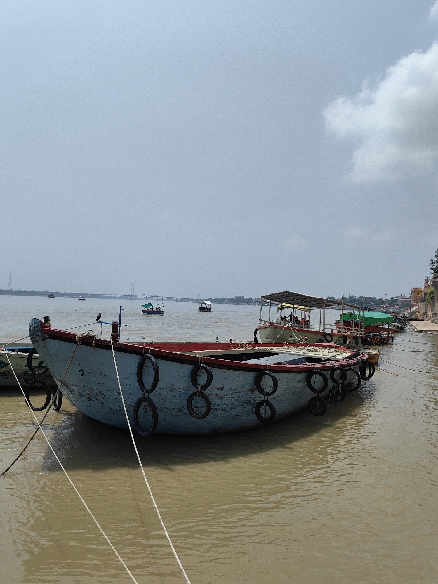 A fleet of boats are moored on the muddy waters of the Ganges River in Varanasi, India. In the foreground, a large, light blue boat with a red trim is prominently featured. It is adorned with black tires, serving as fenders, along its side. Ropes tie the boat to the shore. Other boats are visible in the middle ground, some with canopies and passengers. In the distance, a large bridge spans the river, with more boats and the cityscape of Varanasi lining the far bank. The sky is overcast with white clouds. The scene conveys a sense of activity and daily life along the sacred river.