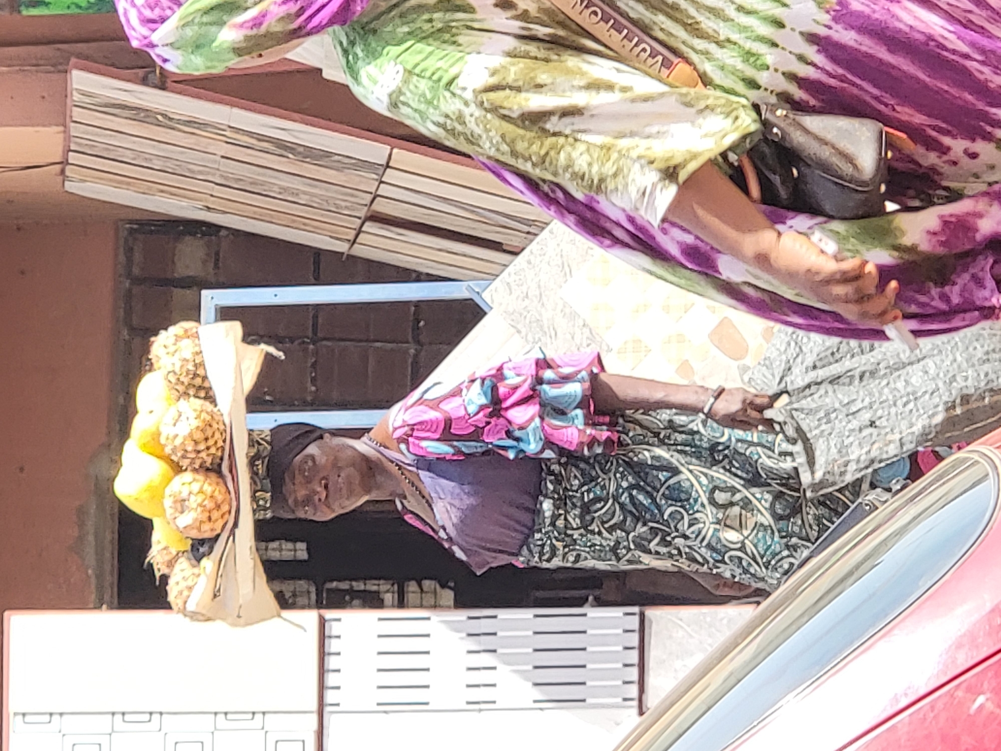 A woman in traditional dress stands on a city street in Maiburiji, Nigeria, selling pineapples and lemons. She wears a dark head covering, a purple-grey short-sleeved top, and a patterned green and blue skirt. She carries a white plastic bag filled with yellow lemons and pineapples. Another person in patterned green and purple cloth, with a Louis Vuitton purse, is standing in the background. A portion of a red car is visible in the lower right. The background shows buildings, including one with a corrugated metal overhang and a barred window.