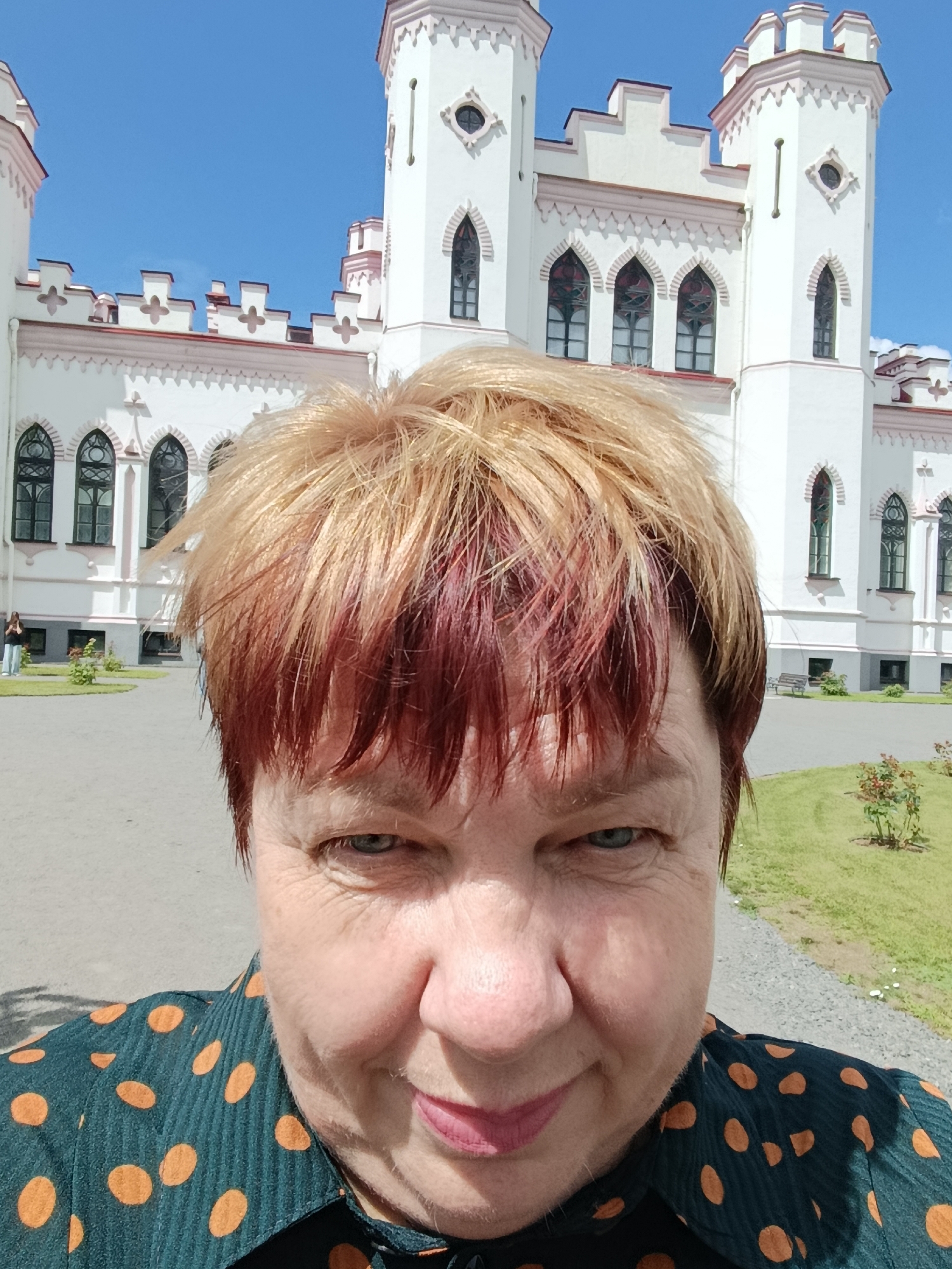 In a selfie taken outdoors, a fair-skinned woman with blonde hair streaked with red smiles at the camera. She is wearing a dark teal shirt with large orange polka dots. Behind her, a white, castle-like building with Gothic architectural elements and a bright blue sky serve as the backdrop. The building features tall towers with decorative battlements and arched windows. The surrounding area is paved with gravel paths, green lawns, and some small rose bushes. The location is the park of the Mir Castle Complex in the city of Mir, Belarus.