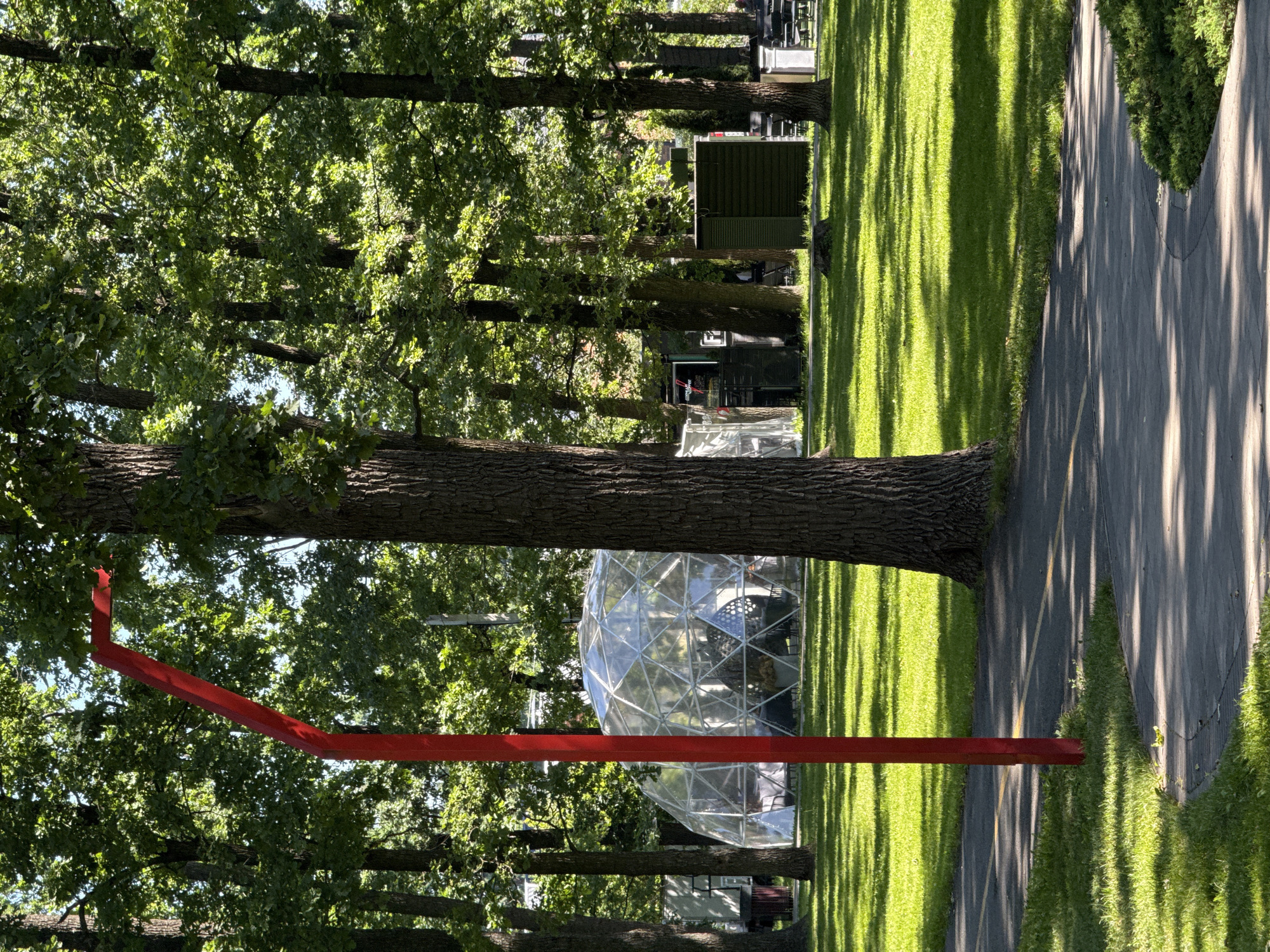 A wide shot captures a park setting on a sunny day, with dappled sunlight illuminating the scene. In the foreground, a paved pathway curves to the right, bordered by lush green grass. A tall, bright red post stands to the left of the path, its angled top disappearing out of frame. Behind the post and pathway, a large geodesic dome, appearing to be made of transparent material, is visible among the trees. The dome is set on a grassy area. Further in the background, several large trees with full green canopies dominate the landscape. Other structures, possibly tents or booths, are also present among the trees. The overall impression is one of an outdoor public space, possibly a venue for an event or gathering, with a mix of natural and man-made elements.