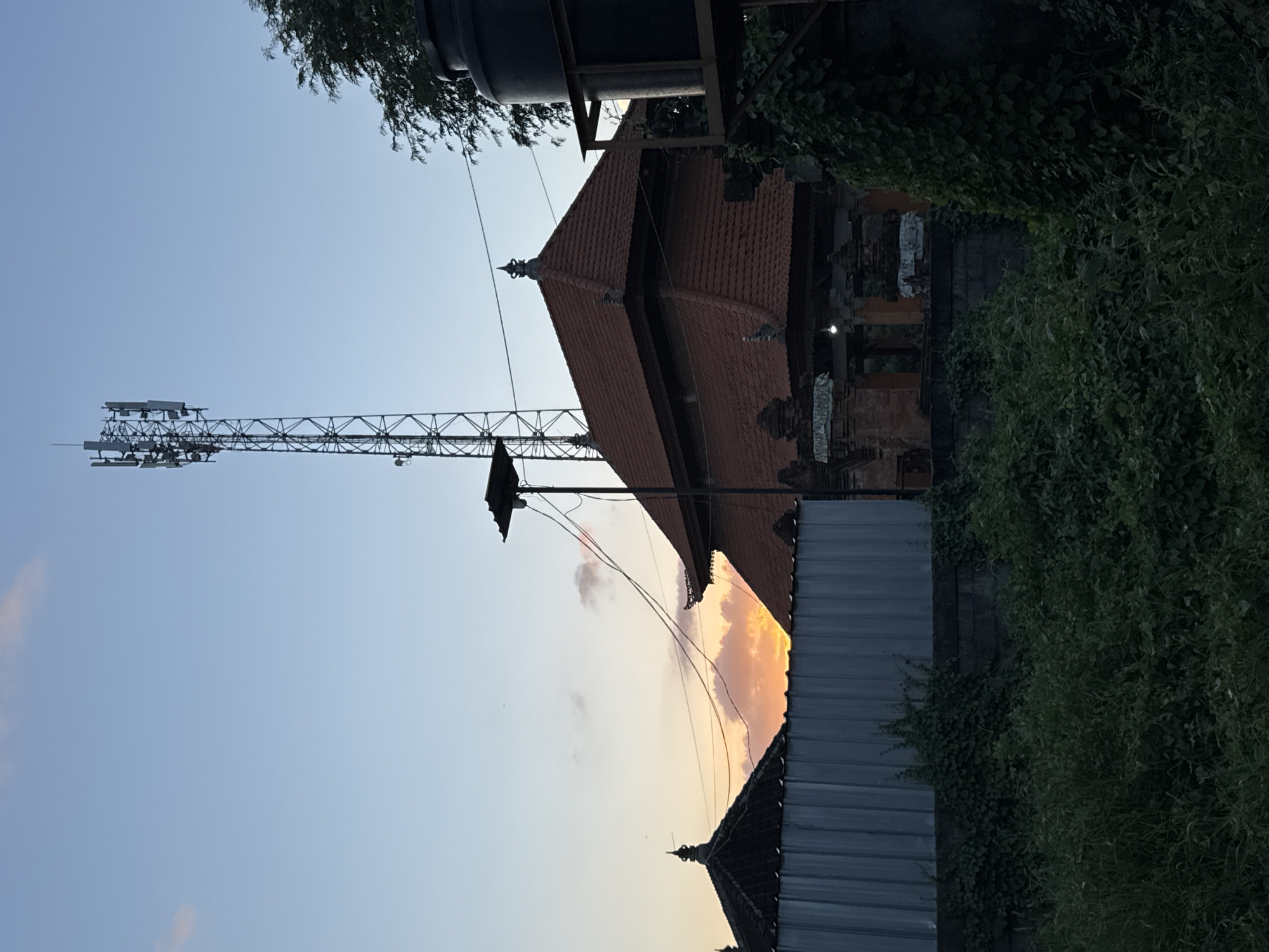 The image shows a view in Batubulan, Indonesia. A cell tower stands against a blue sky with hints of orange from a setting sun. To the right, a traditional Balinese roof with terracotta tiles is visible, along with a water tank atop what appears to be a building or structure. A gray corrugated metal fence runs across the lower right portion of the frame, partially obscured by dense green foliage. Wires are visible stretching between the buildings and the cell tower.