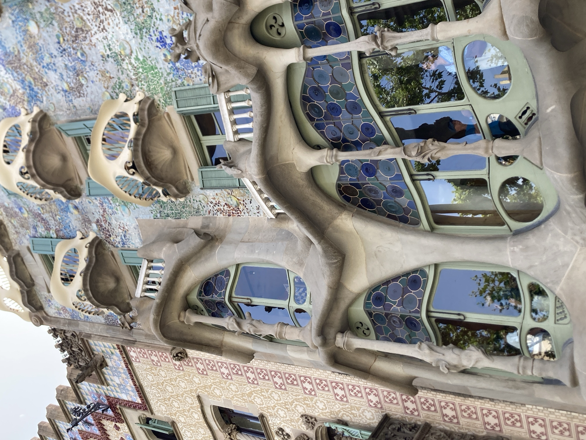 The image shows a close-up, low-angle exterior view of Casa Batlló, a building located on Passeig de Gràcia in Barcelona, Spain. The architecture is characterized by its organic, undulating forms and vibrant, mosaic-like facade. The right side of the building features a mosaic of colorful, irregular tiles in shades of blue, green, and white, interspersed with circular glass elements. The balconies, made of a creamy, bone-like material, have openings that resemble skulls. Below these balconies are large, curved windows framed in a light green hue, with stained glass panels featuring circular blue and green patterns. The left side of the building displays a different facade pattern, with beige tiles decorated with repeating red and cream geometric motifs. Architectural elements like columns with carved details, ornate window frames, and shutters painted a light turquoise are visible. Reflections of trees and the sky are visible in the windows, and two people can be seen through the glass. One person, wearing a blue shirt, is looking outwards, while another, wearing a light blue patterned garment, is looking towards the right. The overall impression is one of intricate and unconventional architectural design.
