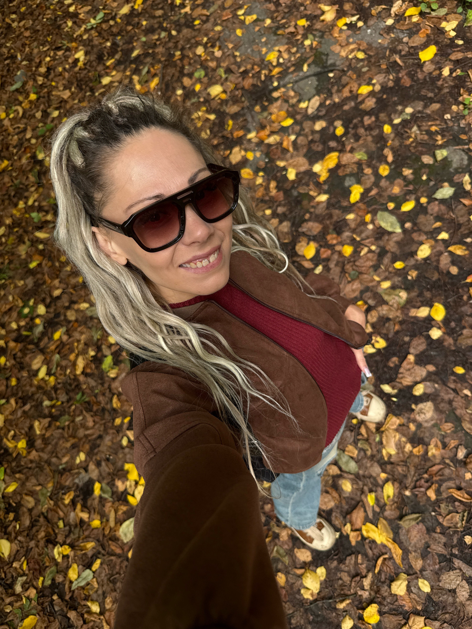 A woman with blonde dreadlocks, wearing oversized dark sunglasses, a maroon top, a brown jacket, and blue jeans, is taking a selfie outdoors. She is standing on a path covered in fallen brown and yellow leaves, typical of autumn. The ground is wet, suggesting recent rain. The background is a blur of trees and foliage. The location is identified as Rusanovo, Russia.