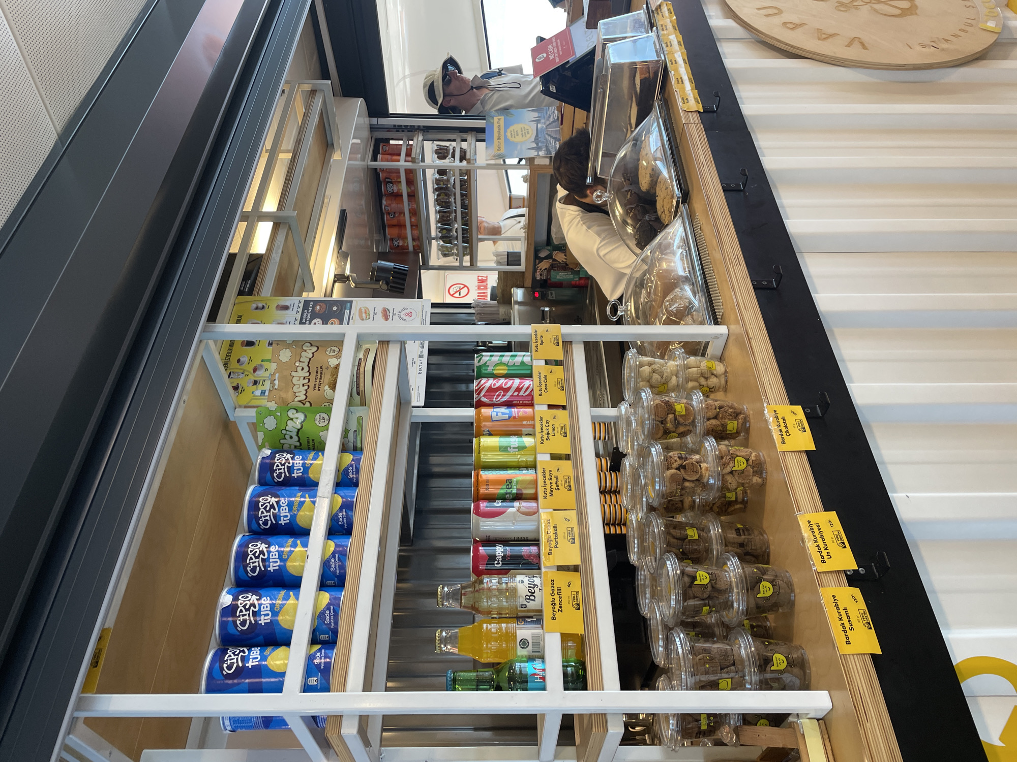 The image shows a street-level kiosk or small shop, likely a food or beverage vendor, with the prominent signage "VAPUR" and "ISTANBUL" suggesting a location in Istanbul, Turkey. The kiosk is constructed from corrugated metal and wood, with open shelving displaying various products. On the top shelf, there are several cans of "Cipso Tube" potato chips and bags of "Pufkins" snack puffs. The middle shelf holds a selection of bottled drinks, including "Hanem" and "Beyoglu Gazoz," along with canned beverages like Cappy and Coca-Cola. Below this, rows of clear plastic containers filled with cookies and baked goods are visible. A man wearing a hat and sunglasses is standing behind the counter, interacting with the scene. Another person is leaning over the counter, partially obscured. The overall environment suggests a busy commercial area, possibly a ferry terminal or a popular tourist spot in Beyoğlu, given the presence of the "Beyoglu Gazoz" label.