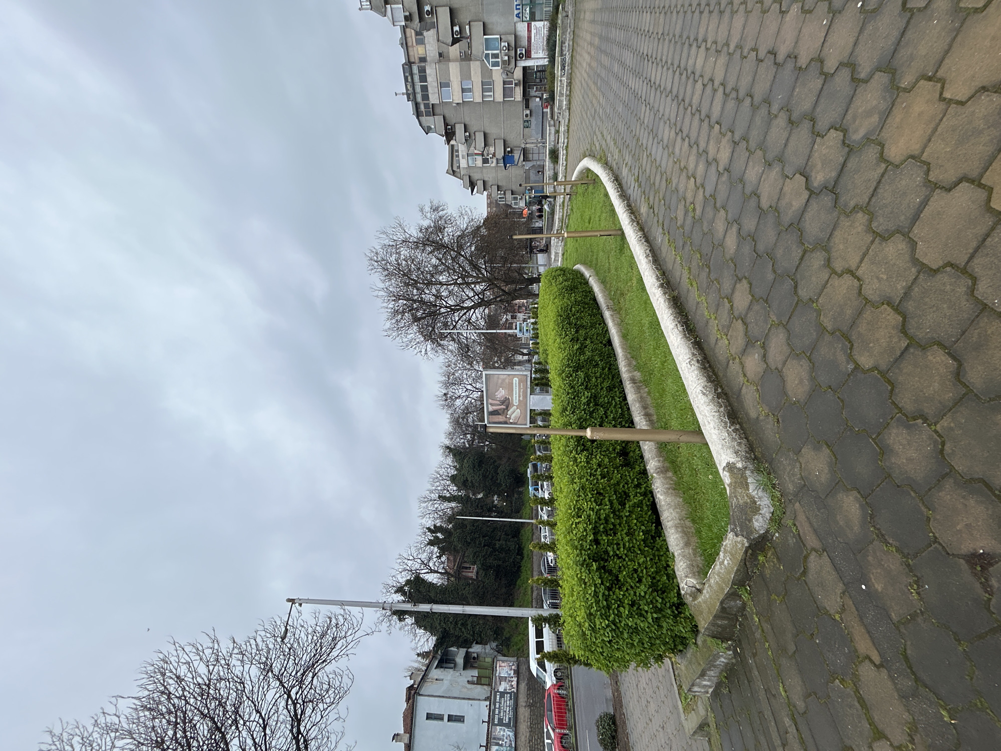 The image depicts a street scene in Plovdiv, Bulgaria. The street is paved with hexagonal interlocking pavers. A curved, waist-high concrete border separates the sidewalk from a narrow strip of grass and shrubs. In the background, there are buildings, trees (some bare), cars parked along the street, and a large advertising billboard. The sky is overcast. The buildings appear to be residential and commercial. There are no visible people actively engaged in actions or interactions in the primary view. The environment is urban, and the overall mood is muted due to the gray sky. A modern residential building with stacked balconies can be seen in the distance.