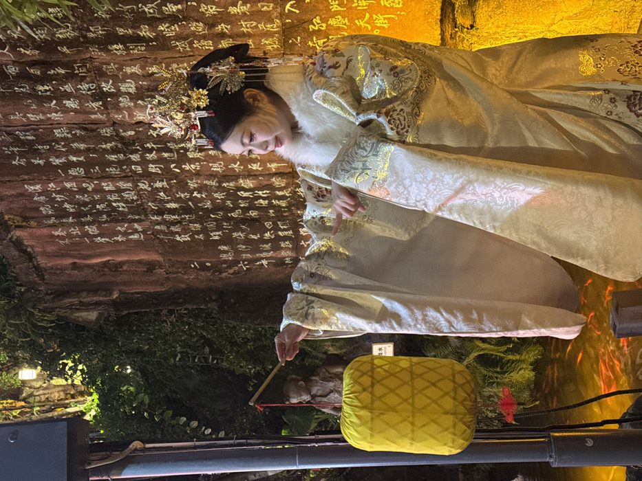 The scene depicts an illuminated outdoor cultural setting, likely within Yuzhong District, Chongqing, China. A single adult female is positioned centrally, adorned in elaborate traditional Chinese performance attire: a gold-patterned brocade robe featuring a white fur collar and an ornate gold-toned headdress with red accents. Her left arm is extended forward, holding a slender wooden stick pointed towards a rectangular yellow fabric lantern with a criss-cross pattern, suspended from a dark pole on the left. Her right hand's index finger also points towards the lantern. The woman exhibits facial makeup including glitter on her eyelids. Dominating the background is a large, reddish-brown rock face, brightly illuminated by a warm light, covered extensively with vertical lines of carved or painted Chinese characters. To the left of the rock face, dense green foliage is visible, partially obscuring a small, dark-colored statue of a child. Below, a pond or water feature is filled with numerous orange-gold fish. A black speaker cabinet is partially visible in the upper left corner of the frame. The ambient lighting indicates evening or nighttime conditions.
