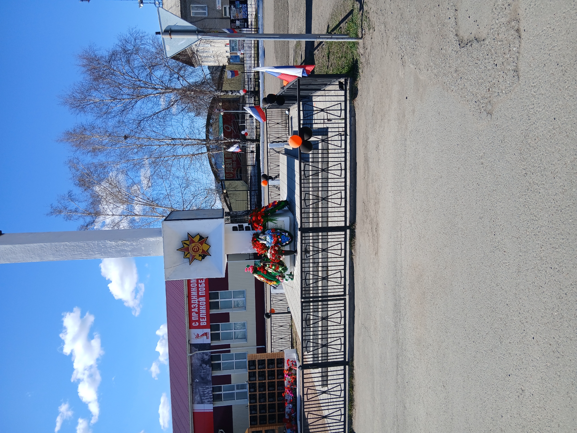 The image shows a war memorial in an urban setting. The memorial is decorated with wreaths and Russian flags. There are black and orange balloons attached to the fence. A building with a banner is visible in the background. The environment is a paved area, possibly a street or a square, under a blue sky with some clouds. The location is in the city of Unknown, Unknown.