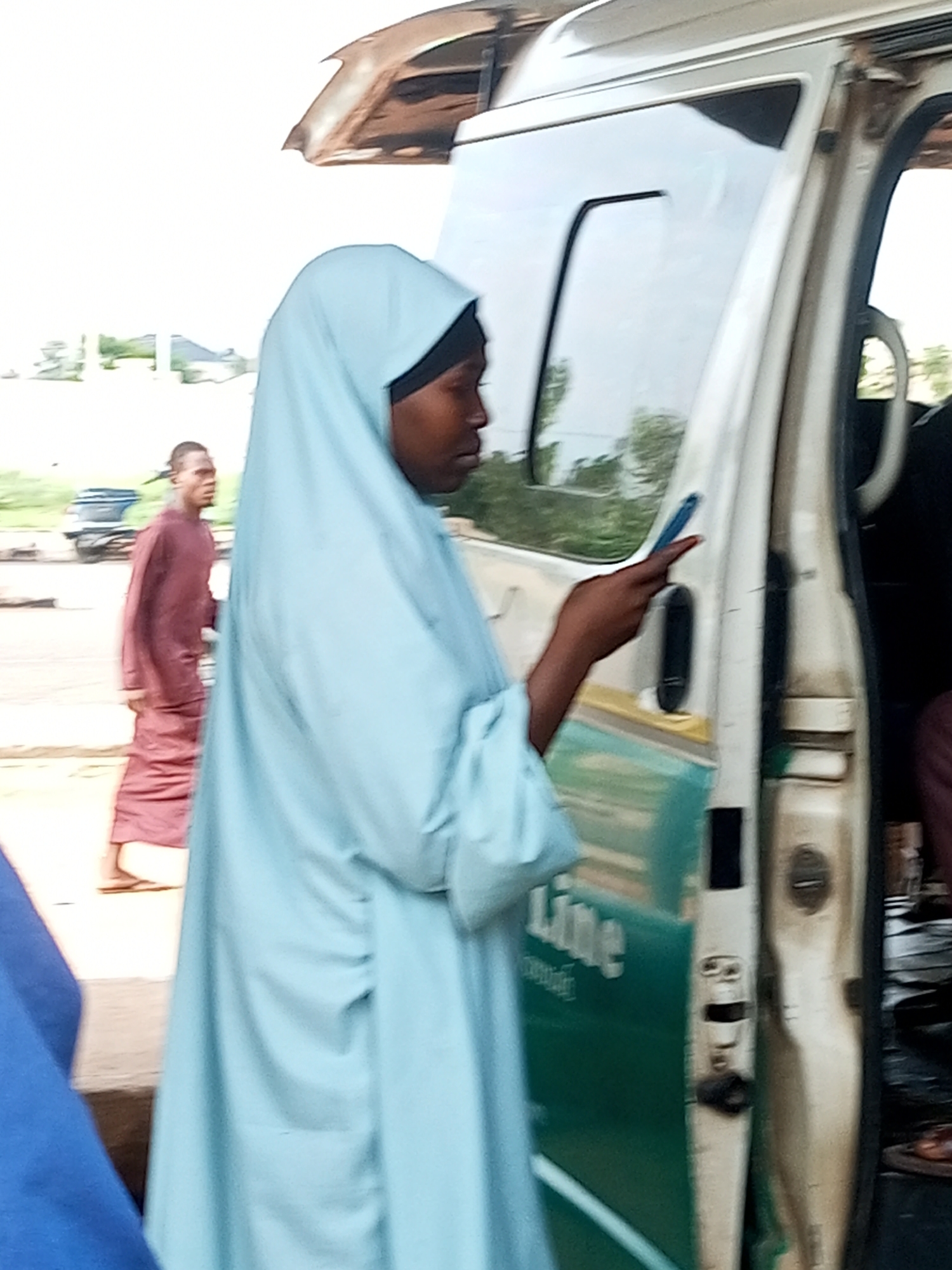 The image shows a woman in a light blue hijab and a light blue abaya standing by the open door of a white van. She is holding a blue object, possibly a phone, in her right hand and pointing it towards the van. Her face is partially visible, and she appears to be looking at the object. In the background, a man in a maroon robe is walking away from the van. The van has a green stripe on its side with the word "Line" visible. The scene appears to be outdoors, possibly at a bus station or roadside stop. The environment suggests a dry climate with some vegetation in the background. This image was taken in Gombe, Nigeria.
