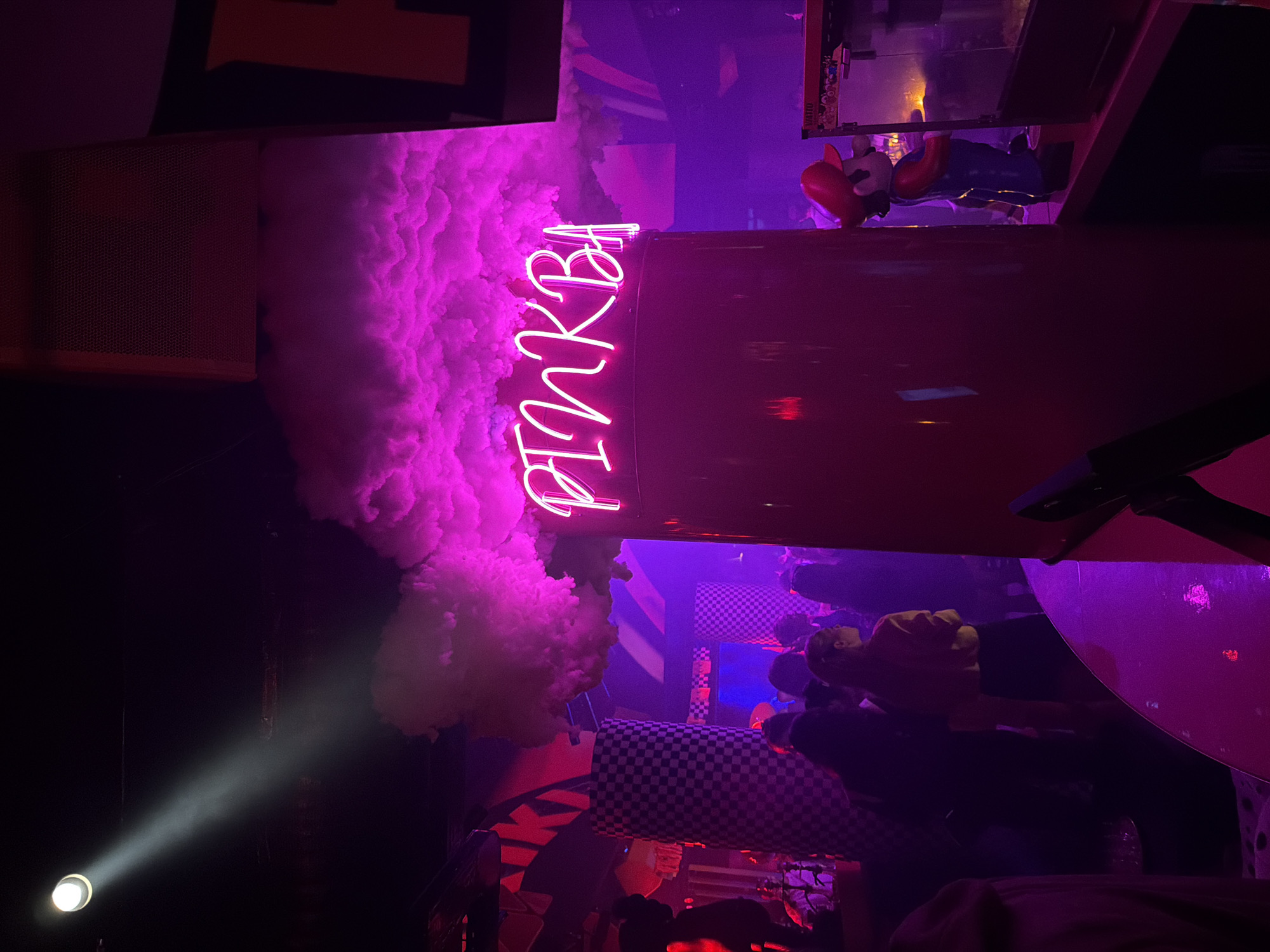 The image depicts the interior of the "Pink Bar" in Moscow, Russia. The scene is dominated by a pink neon sign reading "PINKBA," positioned near a large cloud of artificial smoke. The bar's décor includes checkered patterns and red accents. A figure of Mario, the video game character, is visible on a shelf above the bar. In the lower portion of the image, there are people in the background, partially obscured by shadows. A spotlight is shining from the bottom left corner.