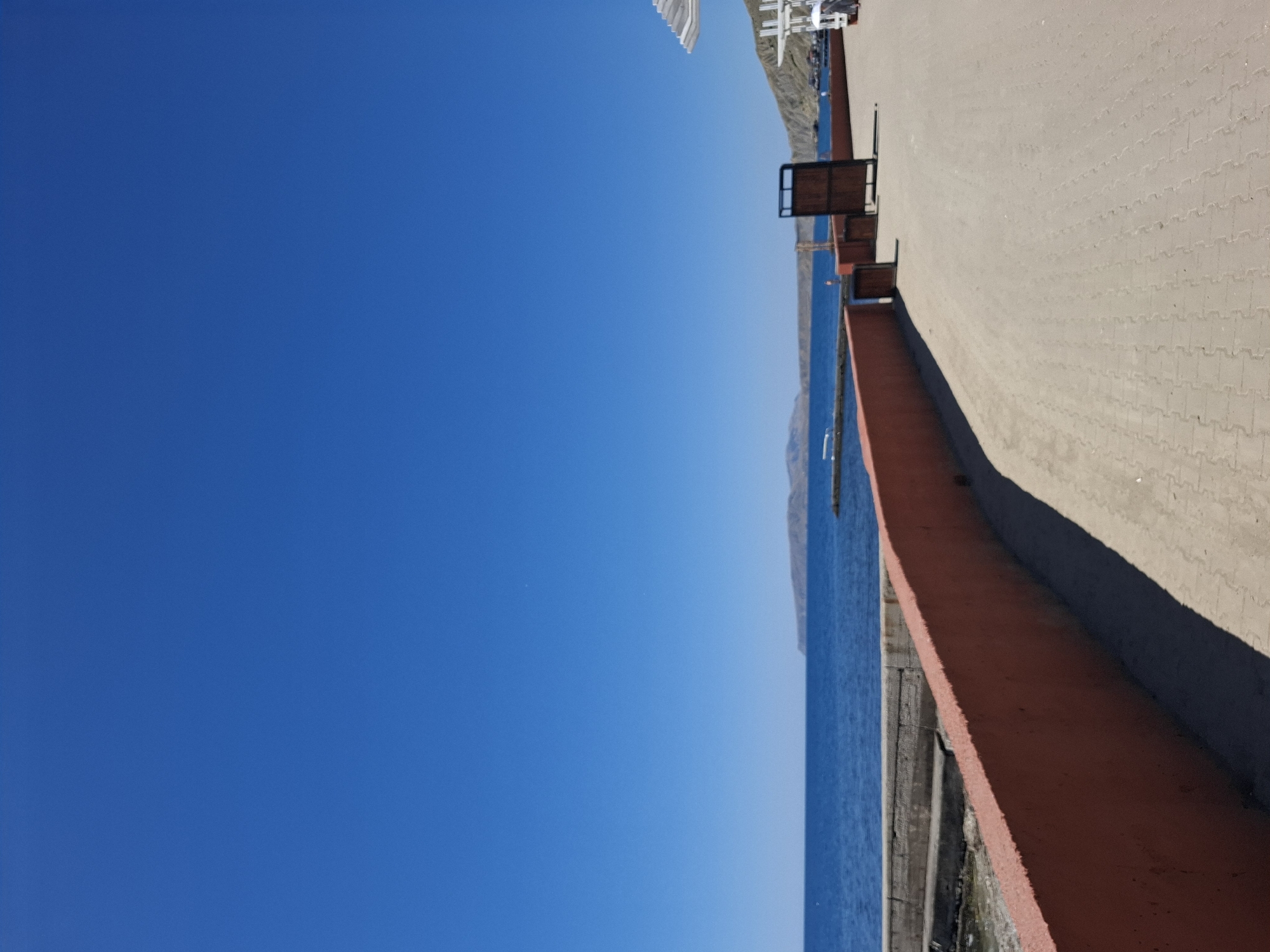 This image depicts a seaside promenade on a bright, clear day. The scene is dominated by a vast expanse of deep blue sky. To the right, a paved walkway with a textured, light-colored surface stretches into the distance, bordered by a low, reddish-orange wall. Beyond the wall, the calm, blue sea meets a distant, hazy shoreline. In the upper right portion of the image, there are structures that appear to be part of a waterfront area, possibly including seating or railing elements. The orientation of the image is sideways. The clear blue sky and bright light suggest it is daytime, likely mid-morning or afternoon, with fair weather. The setting appears to be a coastal town or resort area. No people or specific activities are evident, giving the scene a serene and tranquil feel. There is no visible text in the image.