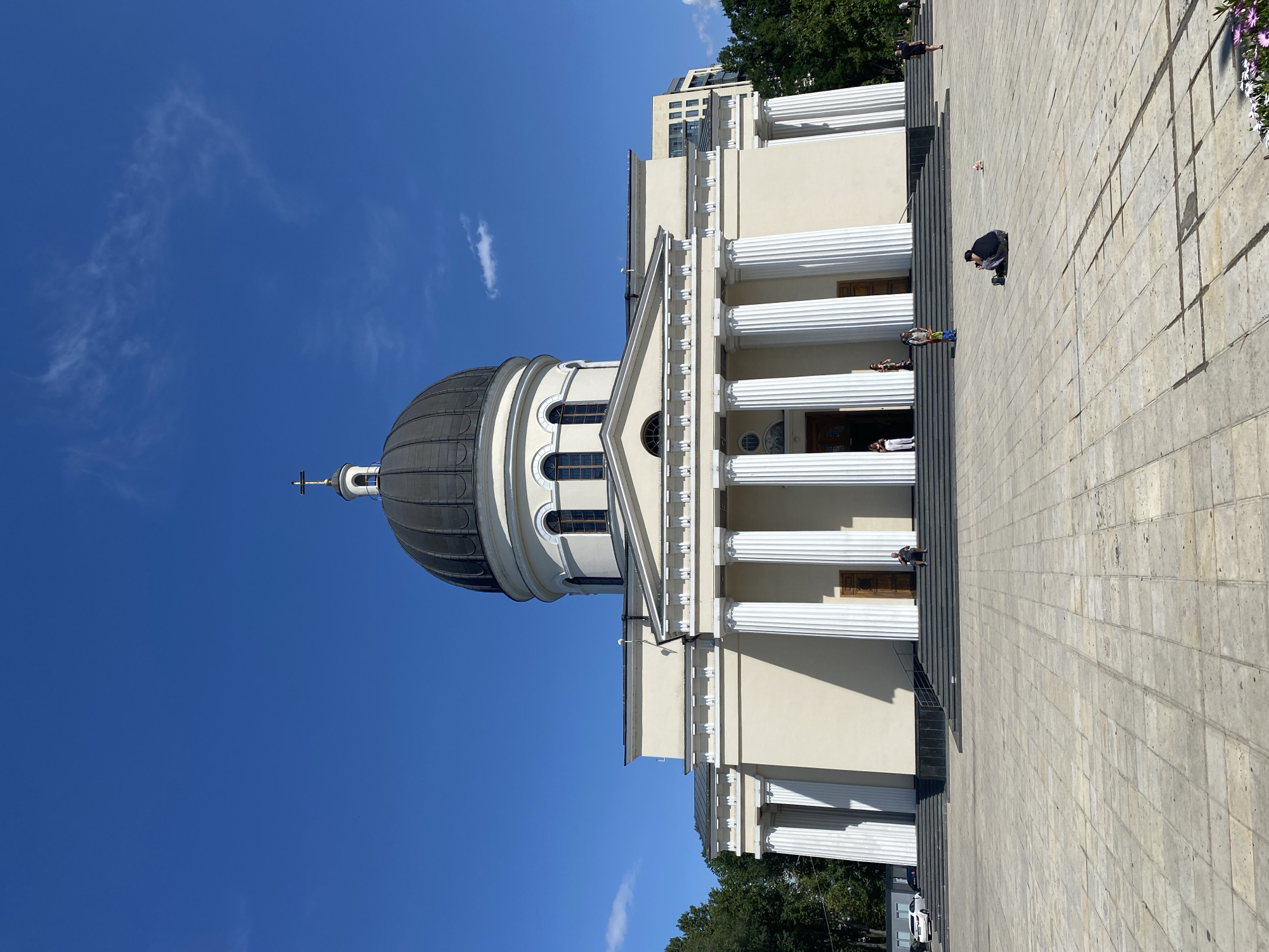 The image shows the Nativity Cathedral in Chișinău, Moldova, under a clear blue sky. The cathedral features a large, dark dome topped with a cross and a prominent portico with white Corinthian columns. In front of the cathedral is a large, paved plaza with some people scattered about. On the left side of the plaza, a white car and a black car are parked. A person is seated on the steps of the cathedral. In the center of the plaza, a person is crouching and appears to be taking a photograph or looking at something on the ground. A small, pink object, possibly a toy, is located a few feet away from the crouching person. The surrounding area is lined with trees.