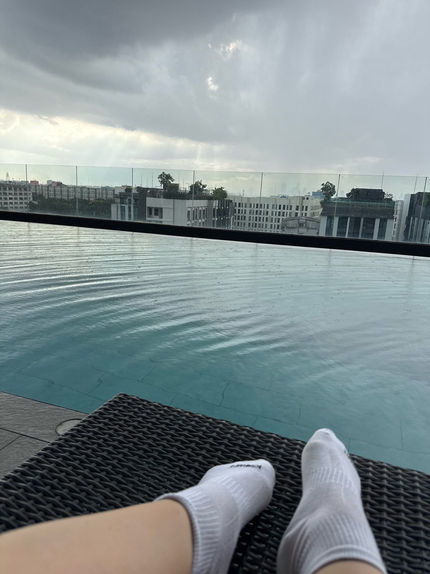 The image depicts a close-up view of a person's feet, clad in white socks, resting on a wicker lounge chair. The chair is situated beside an infinity pool, with the water's surface showing ripples, suggesting light rain. Beyond the pool, a glass barrier separates it from a cityscape. The background features buildings of varying architectural styles, some with intricate facades and others more modern. The sky is overcast with dramatic clouds, hinting at potential rain or a storm. The overall scene conveys a sense of relaxed contemplation, perhaps at a hotel or residential building in the Samrong Nuea Subdistrict Municipality, Thailand.