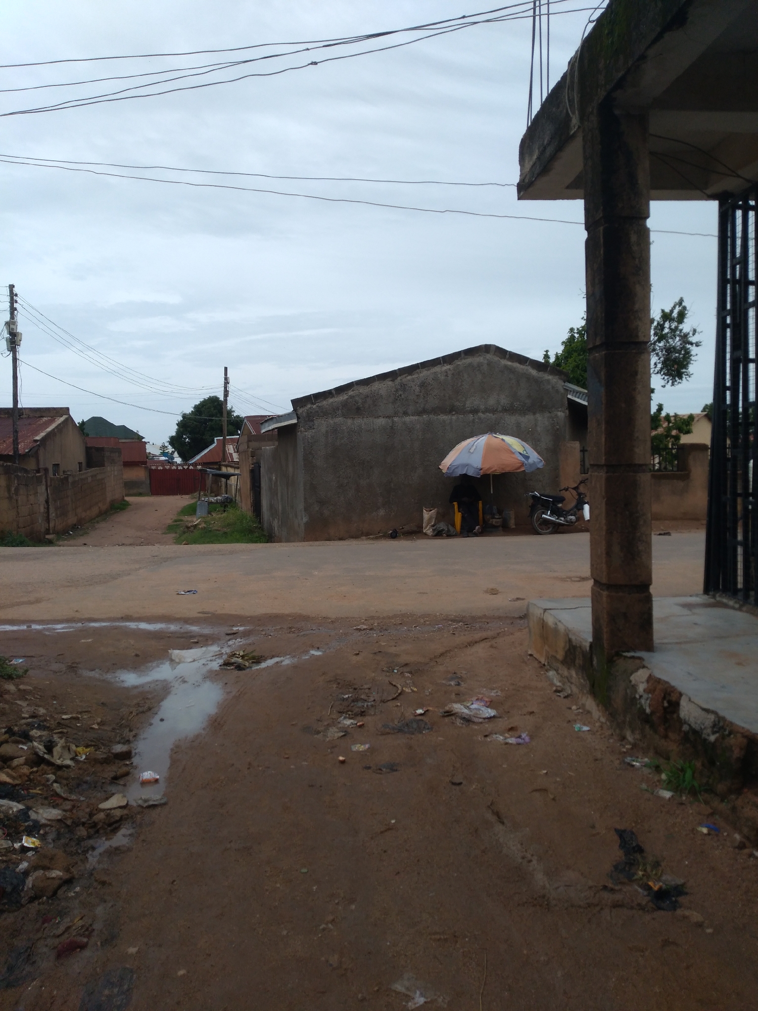 The image depicts a cloudy day in a residential area of Jos, Nigeria, likely a street or alleyway. The foreground is dominated by a muddy, unpaved road with several puddles and scattered debris, including plastic wrappers and rocks. To the right, a concrete structure with a pillar and a partial fence is visible, suggesting a building or entrance. In the midground, a simple, unpainted building with a corrugated iron roof stands. In front of this building, a person is seated under a multicolored umbrella, next to a parked motorcycle. Several sacks are also present. Further into the background, more houses with tin roofs and brick walls line a dirt path that leads to a red metal gate. Overhead, power lines are strung between poles. The overall impression is one of a typical, somewhat unkempt urban environment.