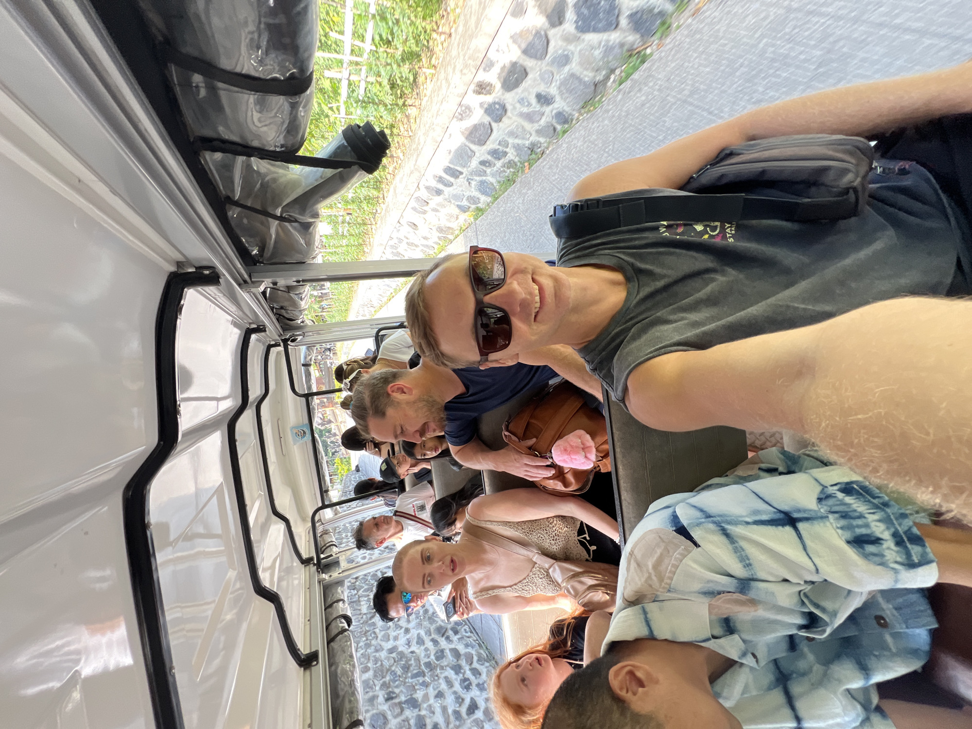 The image captures a group of people riding in an open-air vehicle, possibly a trolley or a tour bus, moving along a paved path. The vehicle has a white roof and appears to be traveling through a green, outdoor environment with stone walls bordering the path.
In the foreground, a man in a dark grey tank top and sunglasses smiles at the camera, holding a dark bag over his shoulder. To his left, another person wearing a blue and white tie-dye shirt is visible, leaning forward.
In the middle ground, several other passengers are seated. A woman in a cream-colored tank top and a light-colored crossbody bag is looking towards the left. Next to her, a man with a dark blue shirt is visible. Further back, other individuals are seated, including a child with red hair.
The environment suggests a sunny day, with greenery and stone structures. The vehicle appears to be in motion, indicated by the slight blur of the background and the way the people are seated. The scene conveys a sense of casual travel or tourism.
No specific details place this scene within the city of Kediri, Indonesia.