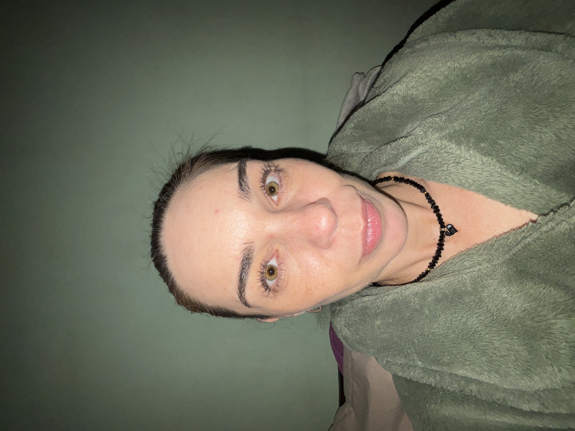 A medium shot photo shows a woman with her hair pulled back and light-colored eyes. She is looking directly at the camera and wearing a green fuzzy robe, a black beaded necklace with a black heart pendant, and earrings. The background is a plain green wall.