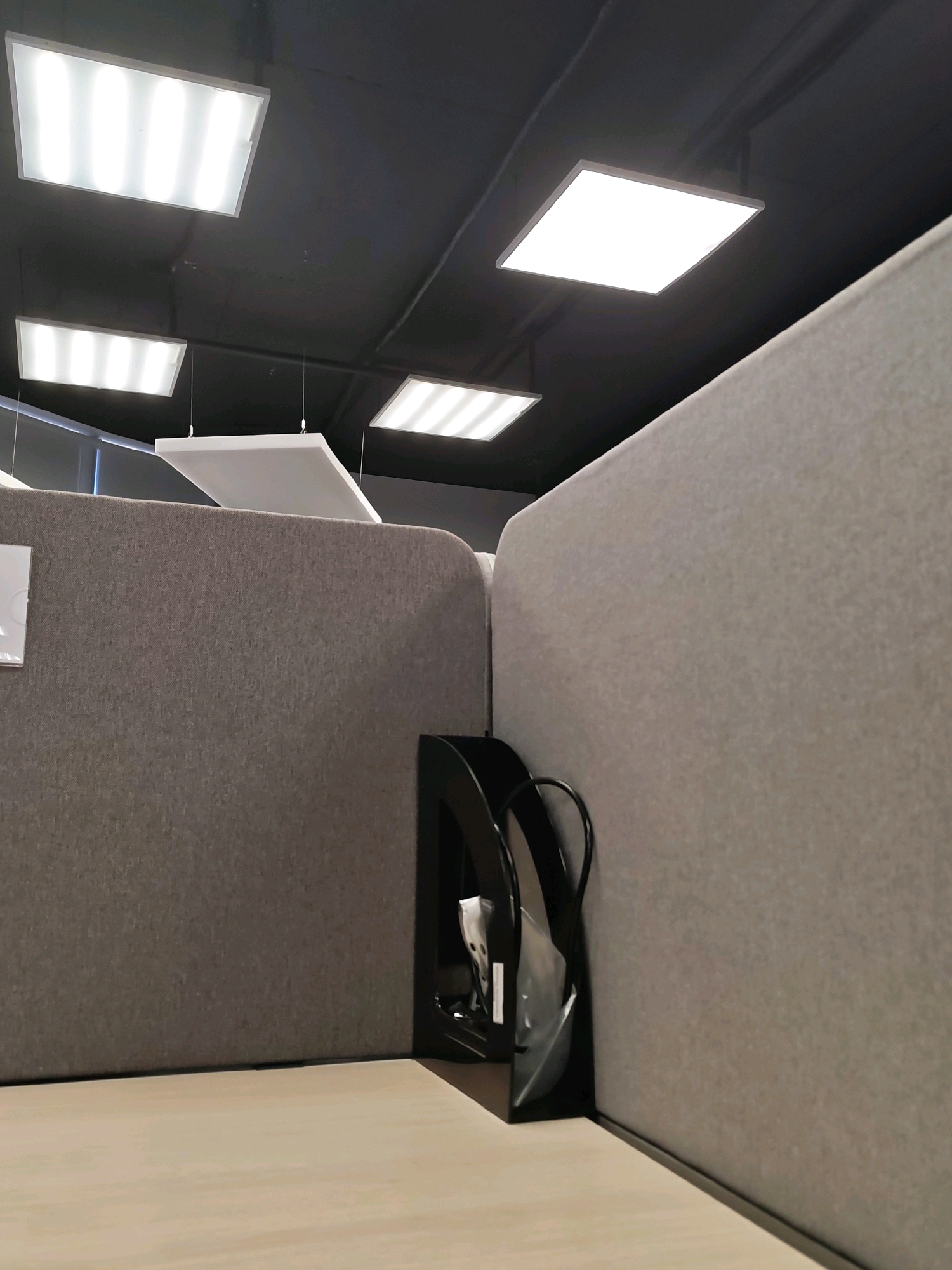 The image depicts an interior view of an office space, likely a cubicle farm, with a focus on the partitions and lighting. The scene is set in Saratov, Russia. The walls of the cubicles are covered in a textured, grey fabric, creating a muted and professional aesthetic. Overhead, modern, rectangular LED light panels are integrated into a dark ceiling, providing bright, even illumination. One panel is square, while others are rectangular. A white, rectangular acoustic panel is suspended from the ceiling by thin wires. In the foreground, a portion of a light-colored desk or tabletop is visible. Attached to one of the cubicle partitions is a black, curved plastic organizer, likely for papers or files, which appears to contain some white items, possibly plastic sleeves or documents. The lighting is bright and artificial, casting minimal shadows, suggesting an indoor, climate-controlled environment. There are no people or animals visible in the image, and no overt actions are taking place. The overall impression is one of a contemporary, functional workspace.