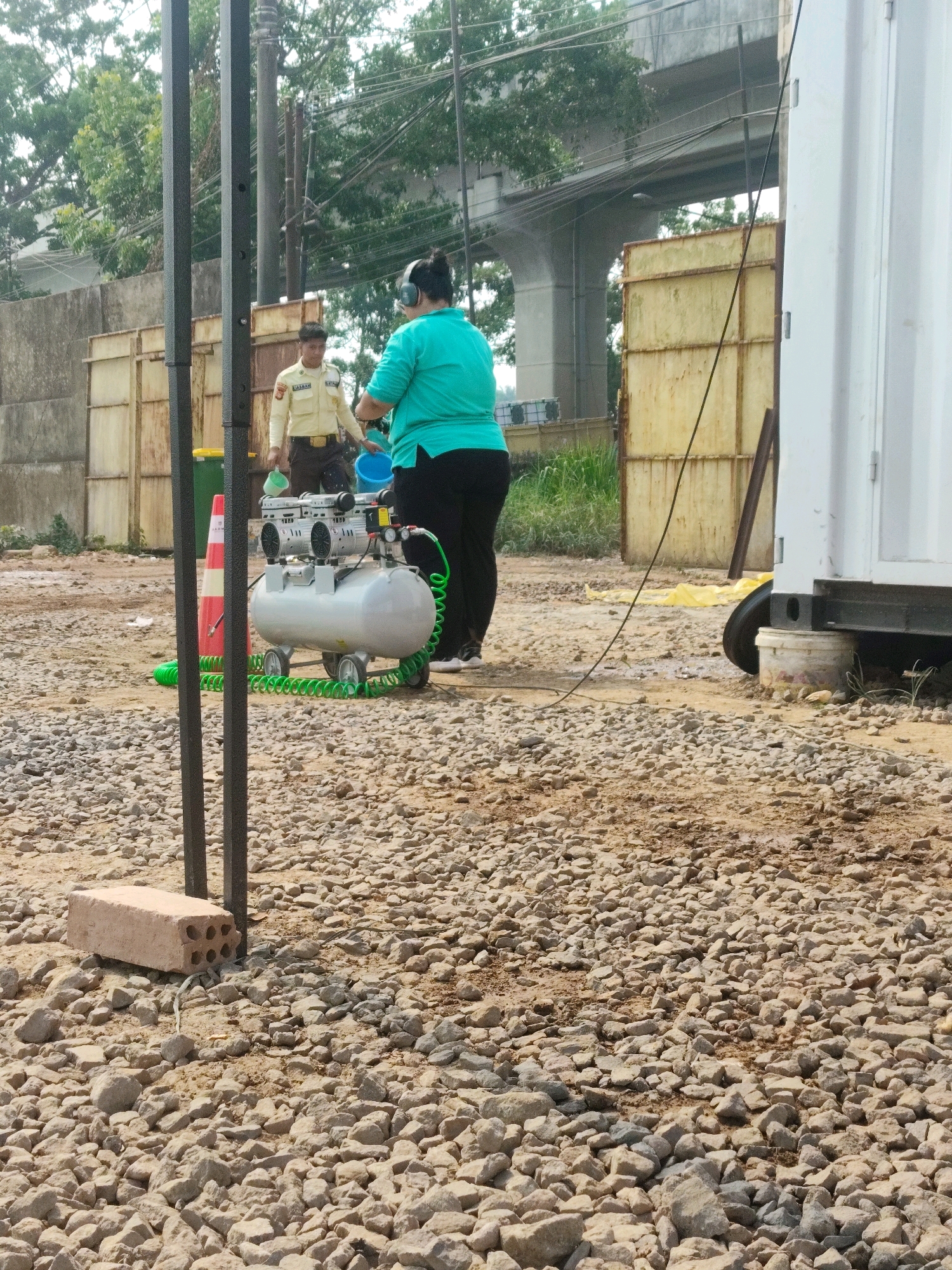 A construction site is depicted in Palembang, Indonesia, with two individuals present. One individual, wearing headphones and a turquoise shirt, is holding a blue bucket, possibly in the process of cleaning or preparing for an activity. The second individual, dressed in a light-colored security uniform with "SAYBAN" written on the back, is holding a green bucket. Between them, a silver air compressor with two motors sits on a wheeled cart, connected by a green coiled hose to a red and white traffic cone. The ground is covered with gravel and dirt. In the background, a concrete overpass and trees are visible, along with a large white shipping container and some yellow metal structures, suggesting ongoing construction or infrastructure work.