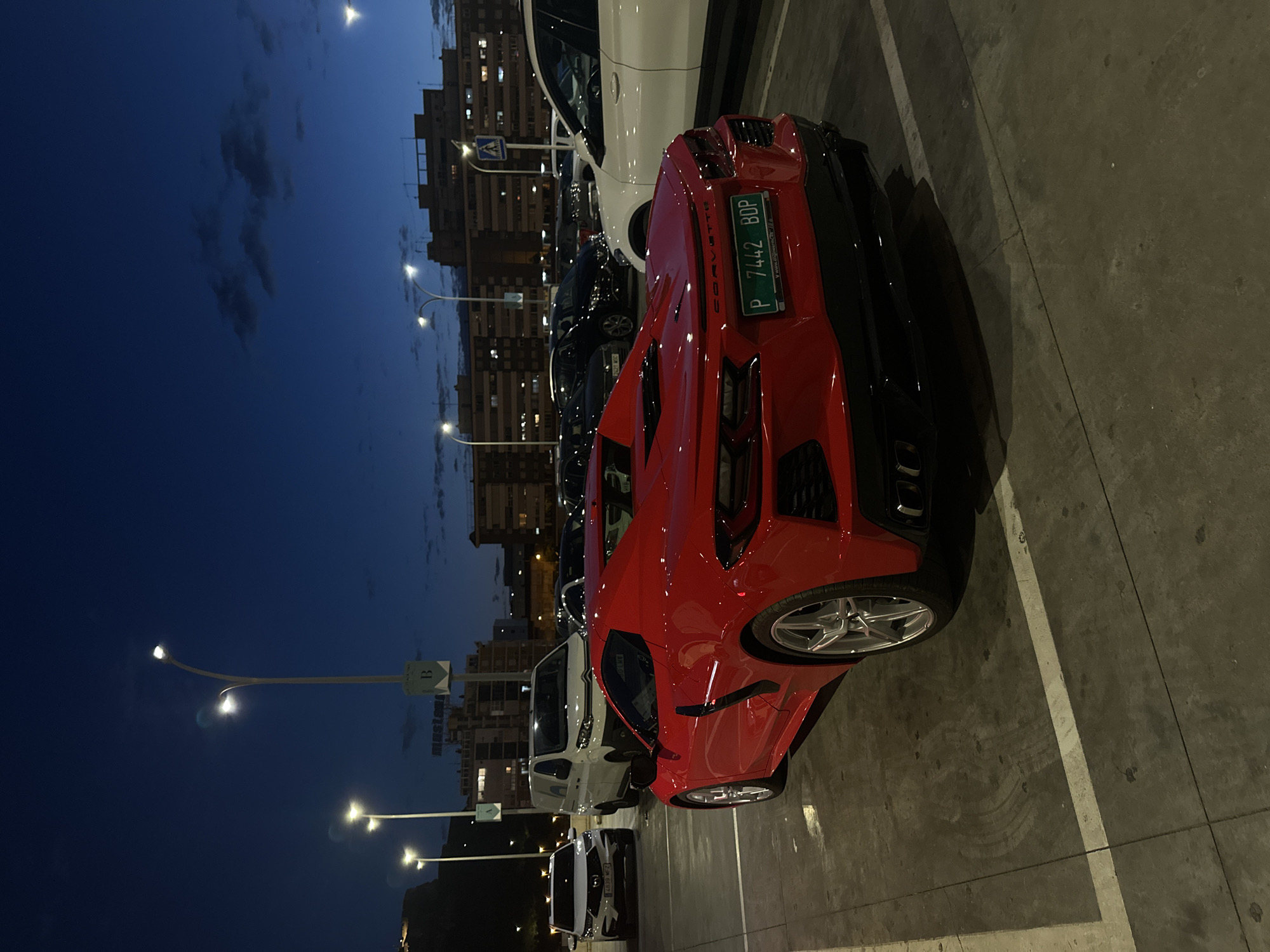 The image shows a red Chevrolet Corvette parked in a parking lot at night. The license plate number of the Corvette is P 7442 BDP. There are other cars parked nearby, including a white car, and streetlights are visible in the background. Buildings are seen in the distance. The location is in the city of Unknown, Unknown.
