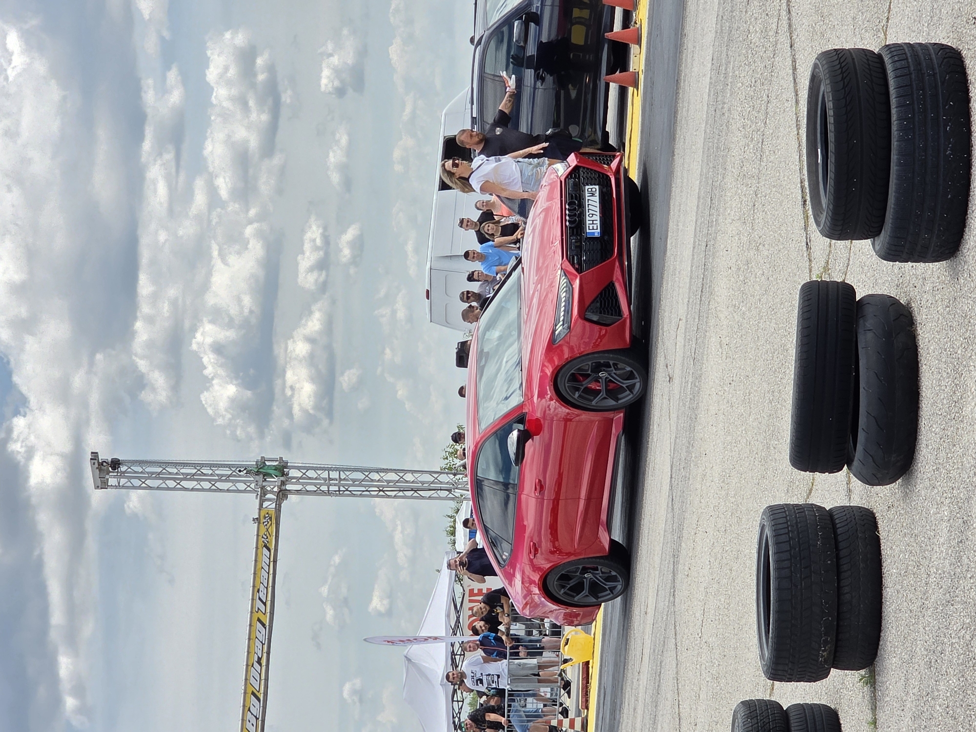 The image depicts a drag race event at the drag strip in Akatsievo, Bulgaria, on a cloudy day. In the foreground, several stacks of used tires are visible on the asphalt track. A red Audi RS 7 Sportback is positioned on the track, facing forward, with its headlights on. To the right of the red car, a black Audi is parked. A group of spectators are gathered on the left side of the track, behind a fence, observing the event. A large metal structure with a banner reading "BIG DRAG TEAM" is visible in the background, along with tents and a white van. Several individuals are standing near the cars, some appearing to be participants or crew members. The overall scene is indicative of a motorsports event, specifically a drag racing competition, taking place in an outdoor venue.