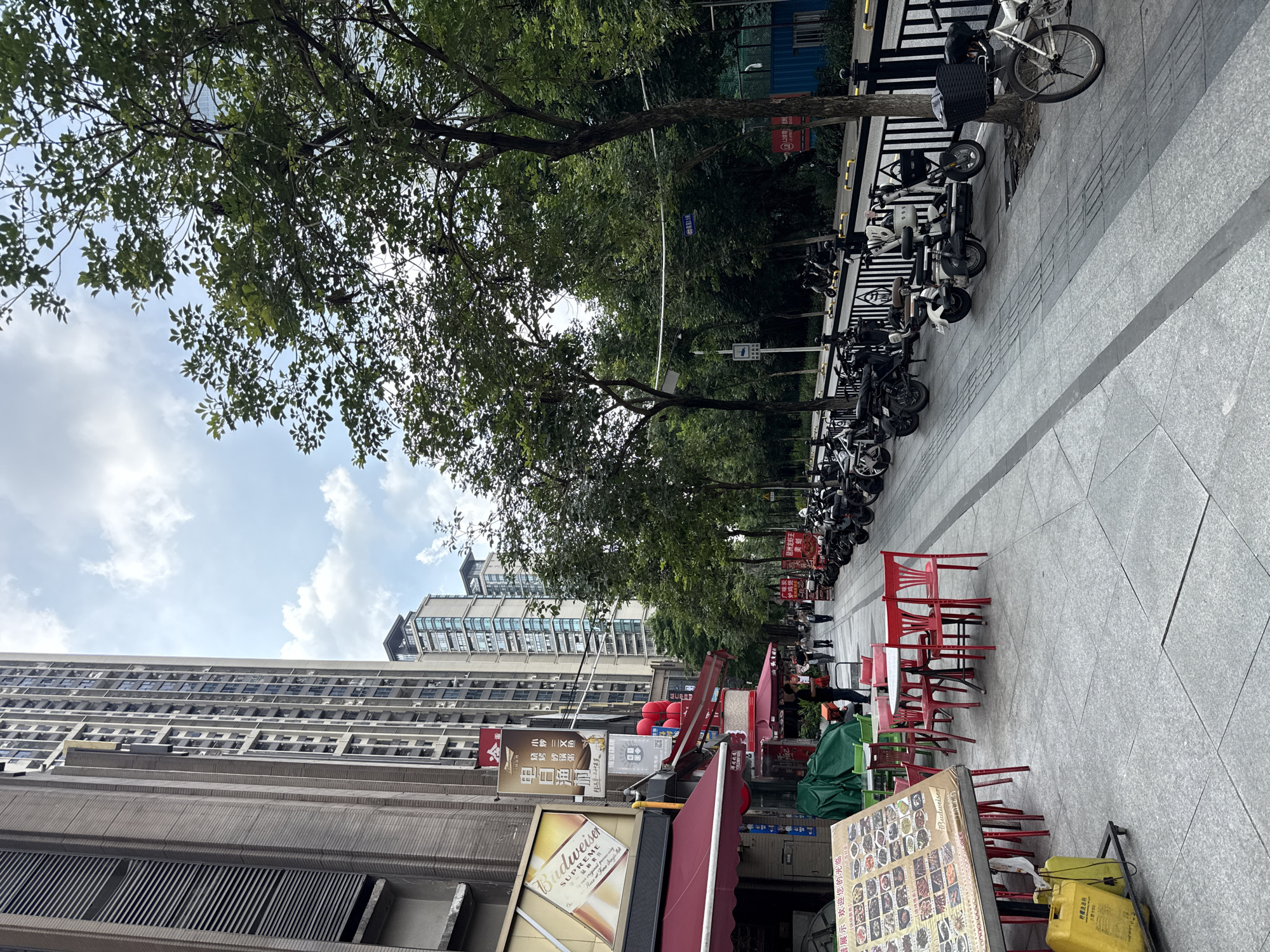 The image shows a sidewalk in Haizhu District, China, with a street lined with trees and buildings. On the left side of the sidewalk, there is a restaurant with red awnings and outdoor seating. A large menu board is set up in front of the restaurant. Beyond the restaurant, several tall apartment buildings rise into the sky. On the right side of the sidewalk, a row of parked motorcycles and scooters is visible. The sidewalk itself is paved with light-colored tiles, and a black metal fence separates it from the street. The street is relatively empty, with only a few people walking in the distance. The sky is partly cloudy.