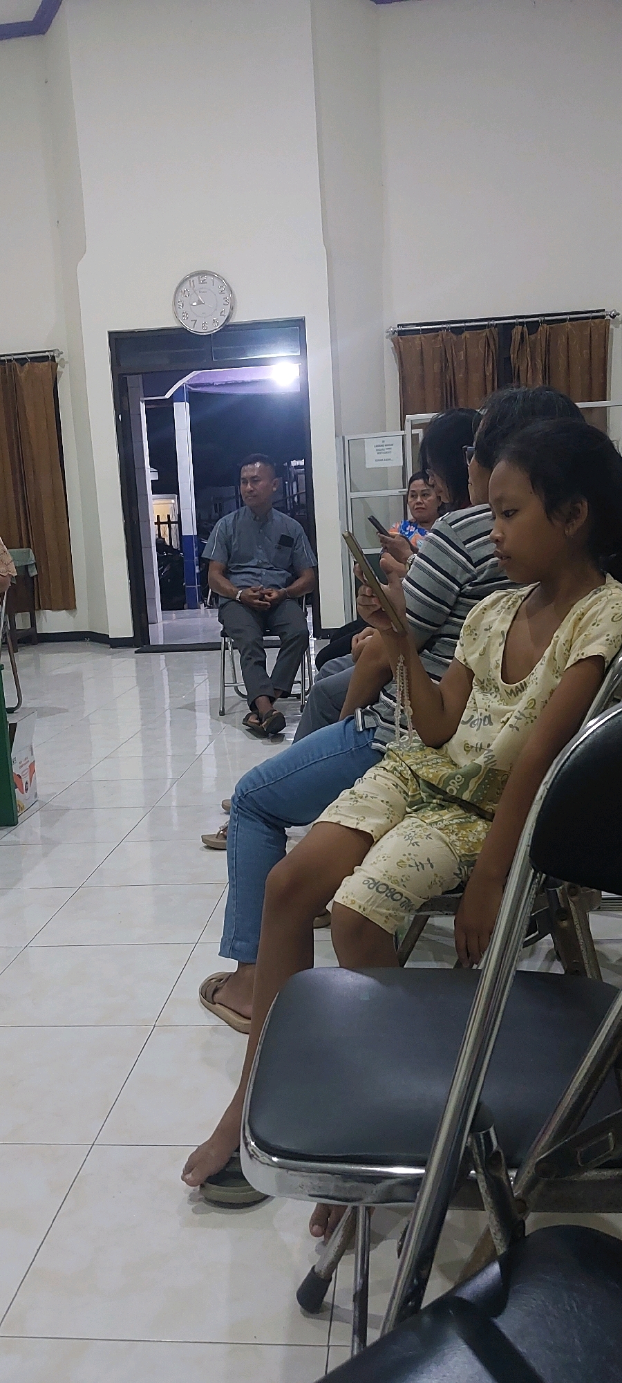 In Cluwak, Indonesia, a man in a grey short-sleeved collared shirt and dark pants sits on a folding chair in what appears to be a waiting area. He is looking towards the left of the frame. In front of him, several people are seated on chairs, facing forward. A young girl in a light yellow top and patterned shorts is in the foreground, holding a book or tablet. Behind her, a person in a striped shirt and blue jeans is also seated. Other individuals are partially visible. The environment includes a tiled floor, white walls, and brown curtains. A clock is mounted high on the wall. The scene suggests a gathering or waiting for an event.
