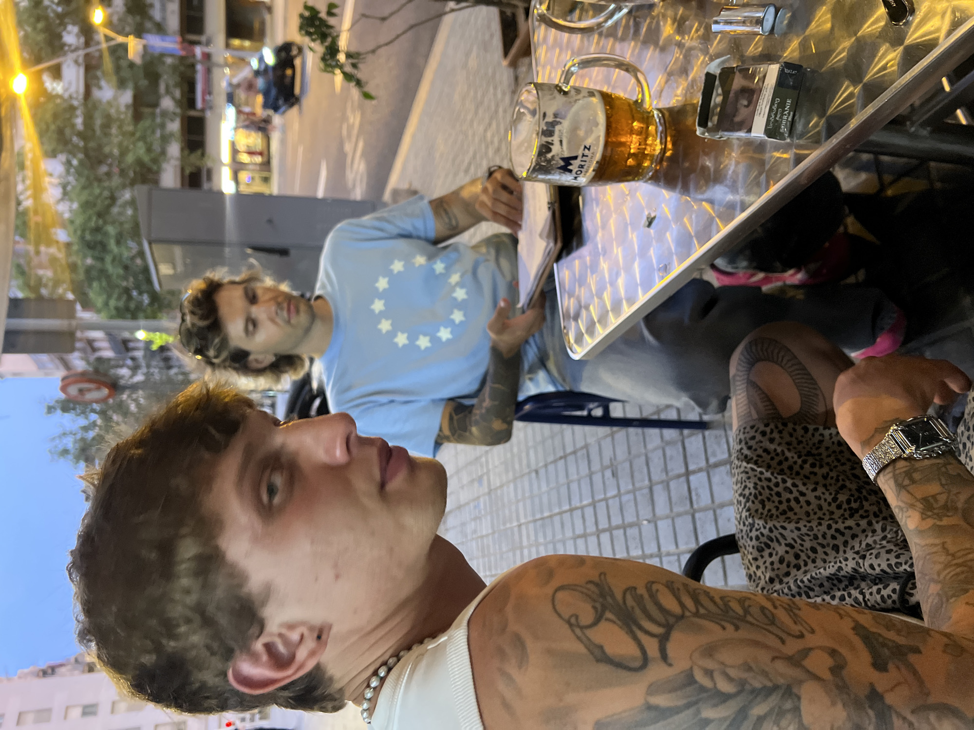 The image depicts two individuals sitting at an outdoor table on a city street in Barcelona, Spain, likely during the evening. The person in the foreground, a young man with light brown, curly hair and visible tattoos on his arms and shoulder, looks towards the camera. He is wearing a white tank top and a pearl necklace. To his right, another man with longer, wavy light brown hair, is seated and looking down at a document. He is wearing a light blue t-shirt with a pattern of white stars resembling the European Union flag.
On the metallic table in front of them sits a glass mug filled with beer, branded with "MORITZ BARCELONA". A pack of cigarettes and a lighter are also present on the table. The background reveals a street scene with buildings, streetlights, and a passing motorcycle, indicating an urban environment. The lighting suggests it is dusk or early evening. The paving stones on the ground indicate a pedestrian area or sidewalk setting.