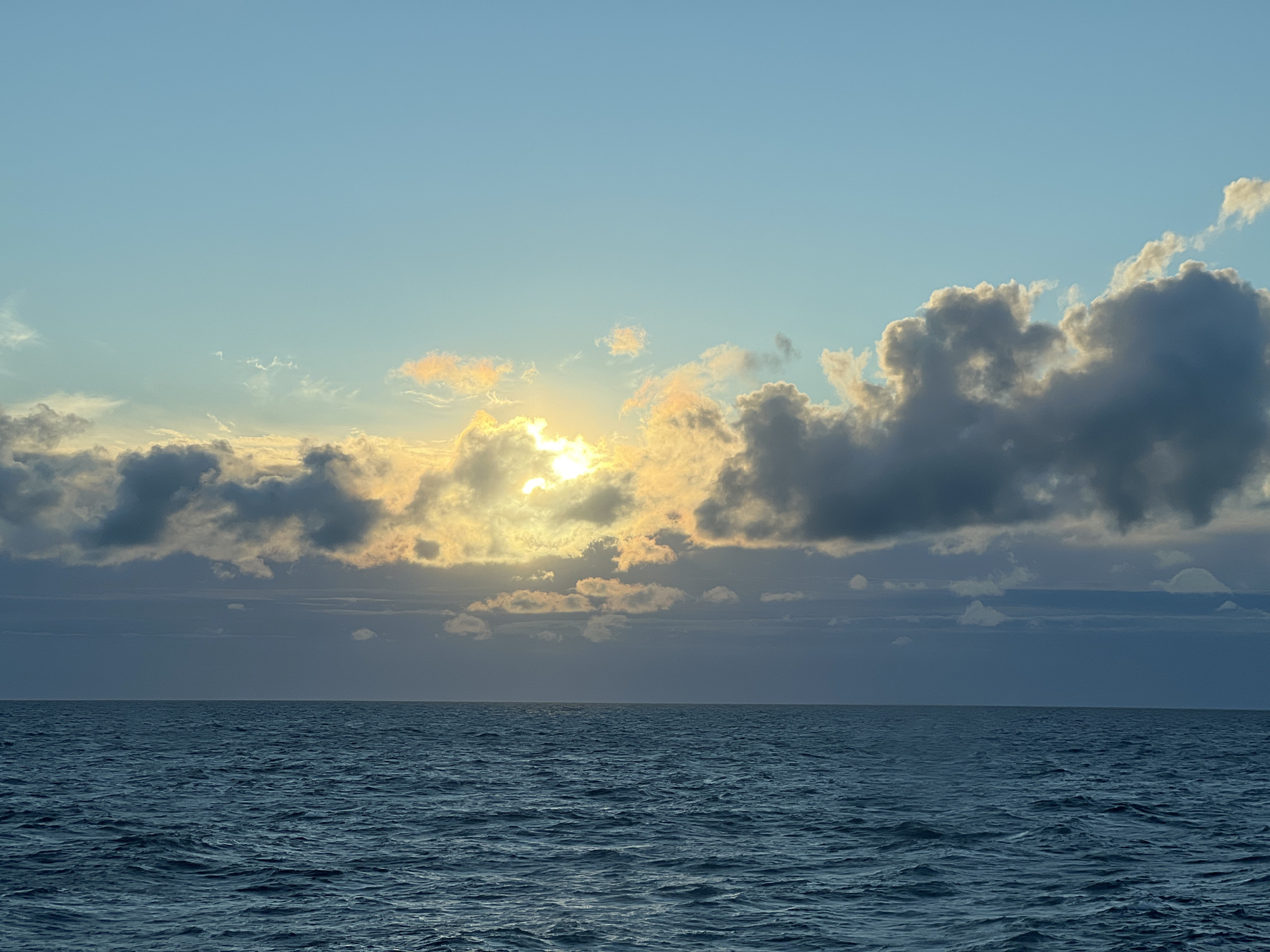 The image shows a wide shot of a seascape at what appears to be sunrise or sunset. The sky is a gradient of light blue near the top, transitioning to softer blues and greys closer to the horizon. Scattered clouds populate the sky; some are dark and heavy, while others are lighter and tinged with orange and yellow from the sun peeking through. The sun itself is partially obscured by clouds, casting a bright, diffused light. Below the sky, a clear horizon line separates the sky from the deep blue, choppy ocean. The water has a textured surface with visible waves and ripples, reflecting the colors of the sky. There are no people, buildings, or specific landmarks visible in the image, making the location in Poland, or anywhere else, indeterminate.