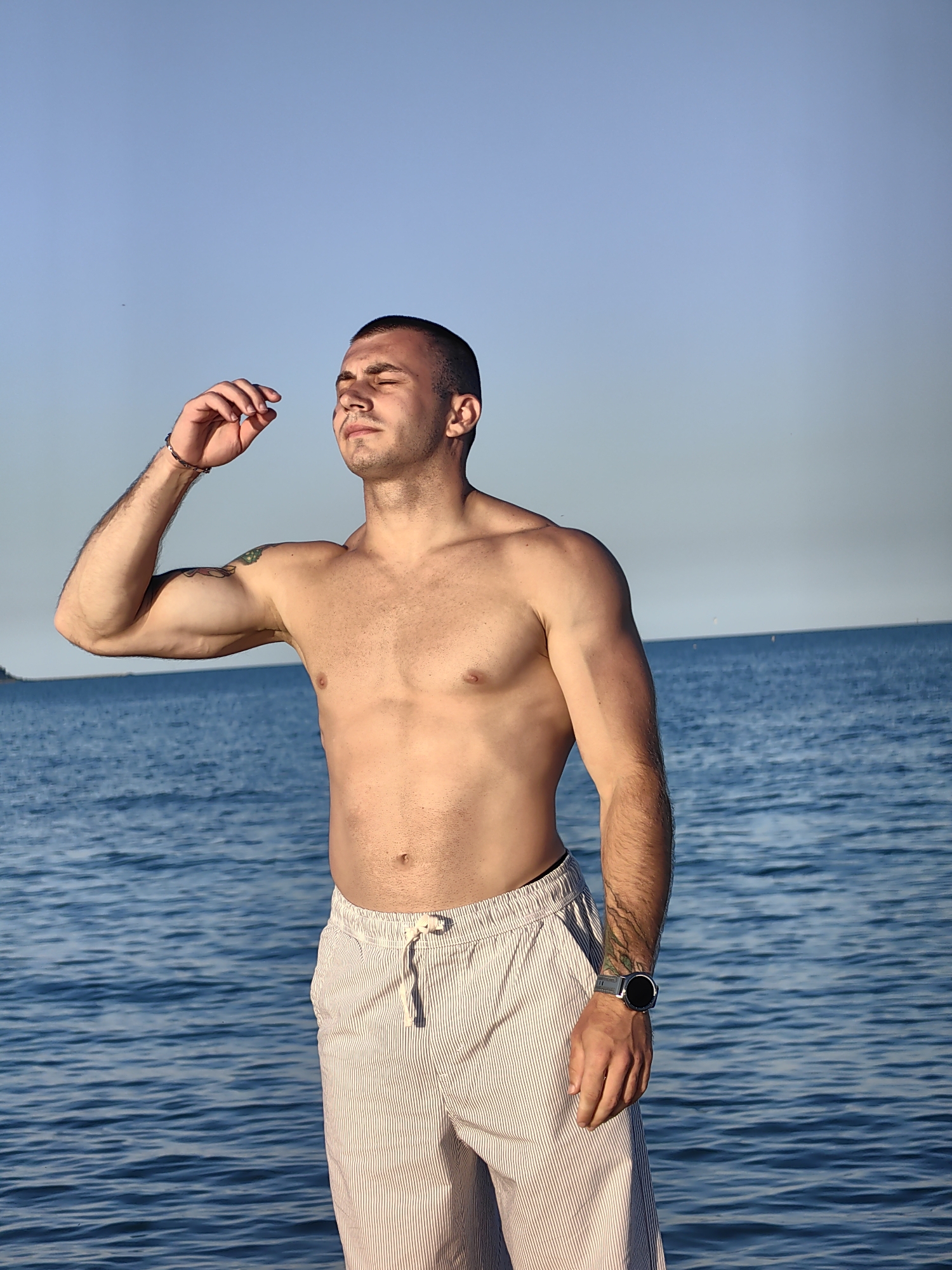 A shirtless man with short hair and closed eyes stands in front of the sea. He wears striped shorts and a watch on his left wrist. His right arm is raised to his face, with his fingers near his temple. The sea is calm, and the sky is clear and blue. The man appears to be meditating or enjoying the moment. There is no indication of the specific location in Varna, Bulgaria.