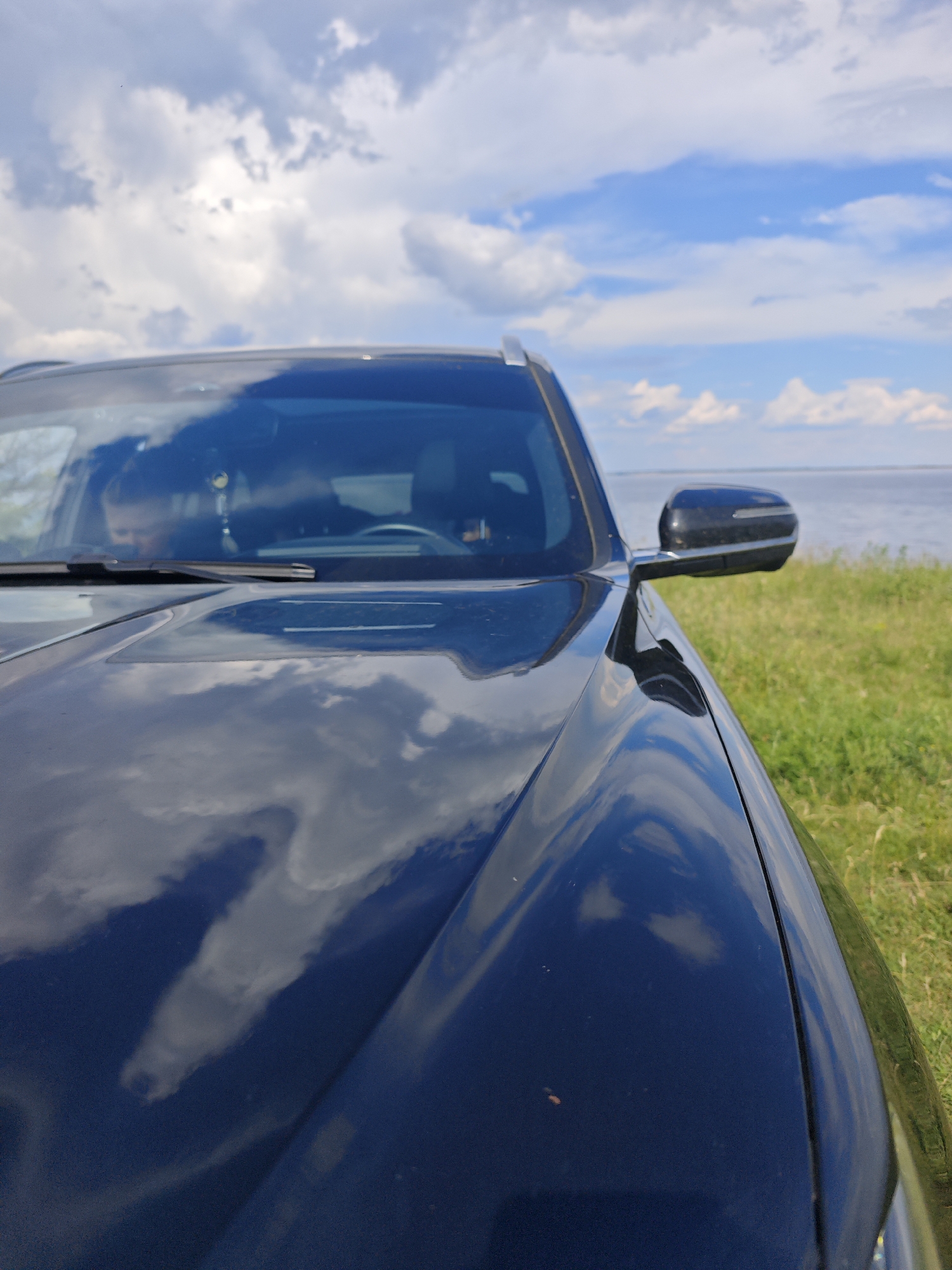 A black car is parked on a grassy area next to a body of water under a cloudy sky. The car's hood reflects the sky and clouds. The scene appears to be in a rural or natural setting, possibly a park or a rest stop. There is no indication of Alekseyevka, Russia.