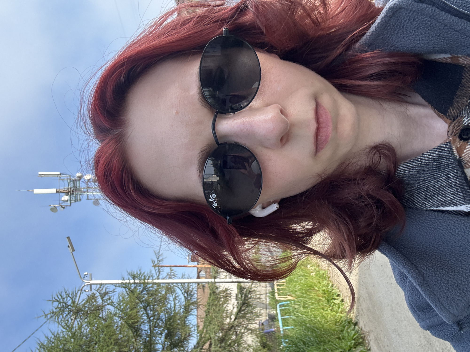 A young woman with dyed red hair and black oval sunglasses is taking a selfie outdoors. She is wearing a grey jacket over a plaid shirt and white earbuds. Behind her, a tree, a street lamp, a cell tower, and a residential building are visible under a clear blue sky. A dirt path is in the foreground. The location is Unknown, Unknown.