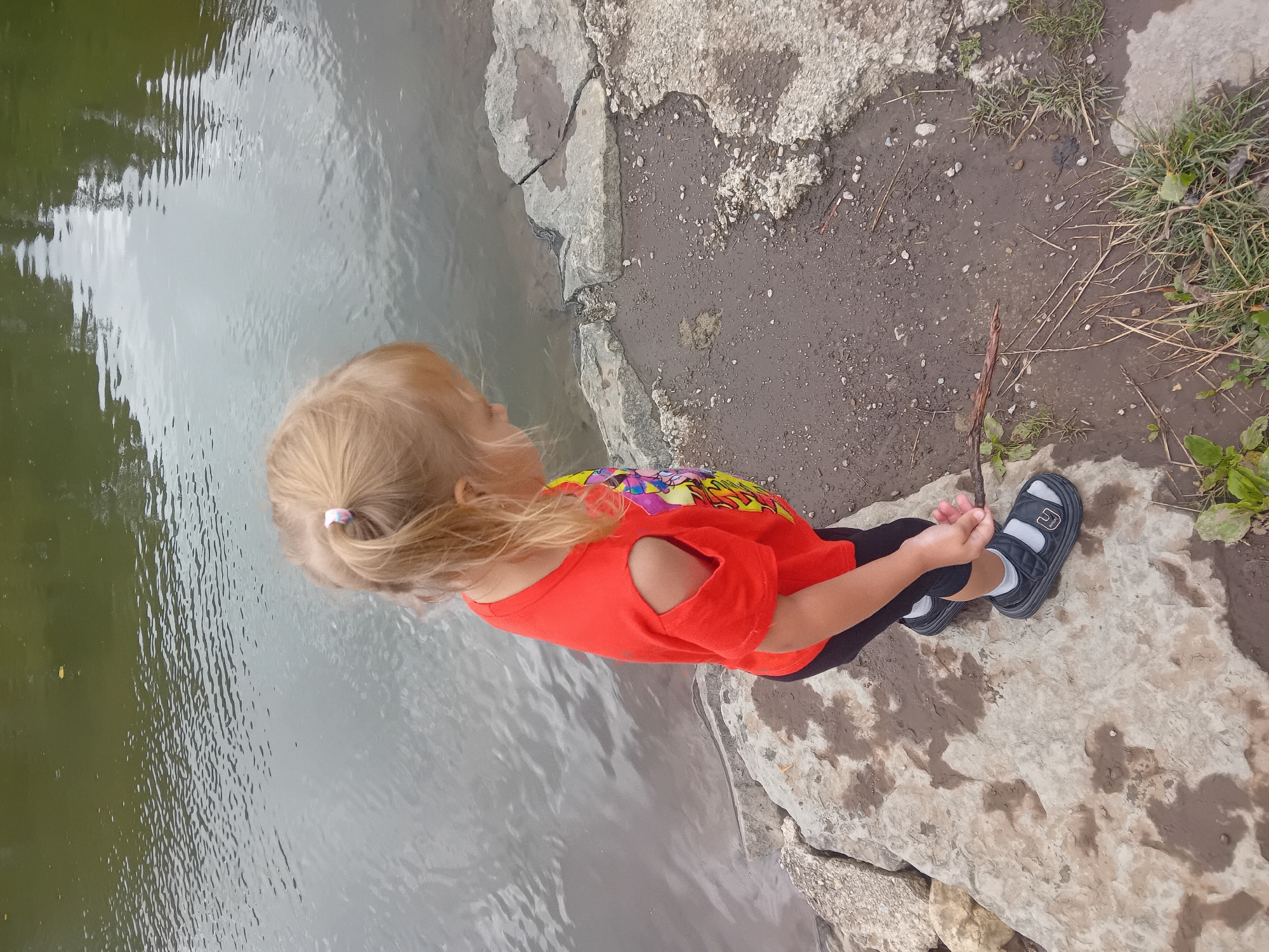 In Habovka, Slovakia, a young blonde child, wearing a bright orange t-shirt with cut-out sleeves and dark leggings, stands by the edge of a body of water. The child is facing away from the camera and looking down towards a stick held in their right hand. The child is wearing black sandals with gray socks. The terrain consists of large, flat rocks bordering the water and a muddy area scattered with small stones and dry grass. The water is calm with ripples and reflections on its surface. The overall scene is outdoors, suggesting a natural or park-like setting.