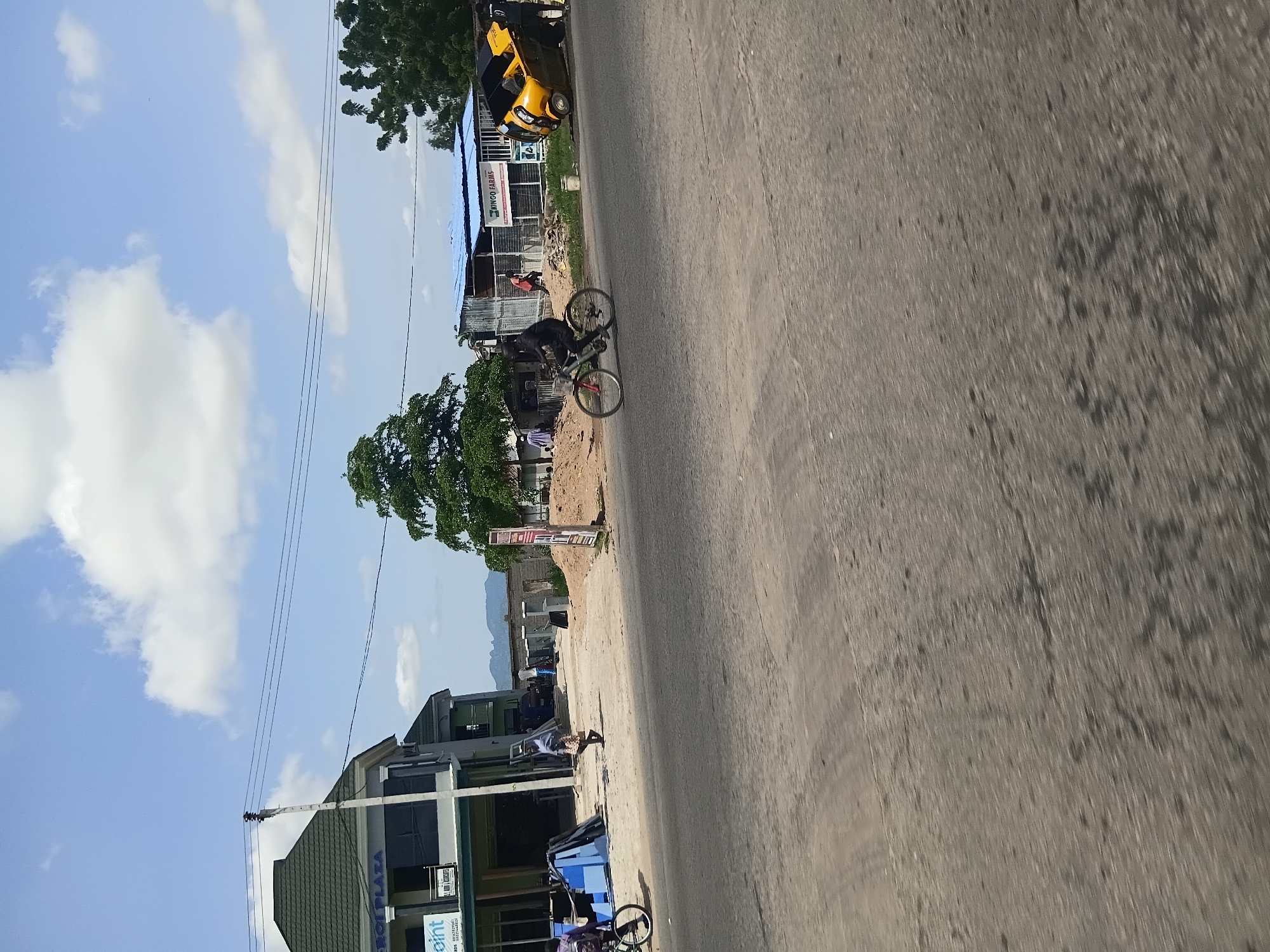 In Jalingo, Nigeria, a wide paved road occupies the foreground of the image, with a significant portion of the frame dedicated to its texture and markings. To the left, a modern, multi-story building with a dark green roof and a sign reading "ERGOPLAZA" is visible. Adjacent to it, another section of the building features a sign with "POINT" on it.
Further down the road, a cyclist in black attire is actively riding a bicycle, their movement blurred by motion. Beyond the cyclist, a yellow three-wheeled vehicle, possibly an auto-rickshaw, has overturned on its side. Two individuals are standing near the overturned vehicle, one in black and another person partially obscured.
In the background, various structures and natural elements are present. There is a smaller building with a blue roof, and further back, trees and a distant mountainous landscape under a partly cloudy sky. A power pole with wires stretches across the scene, and along the side of the road, there are construction materials and piles of debris. People are seen walking in the distance near the buildings. The overall scene depicts a typical urban environment with signs of activity and a minor traffic incident.