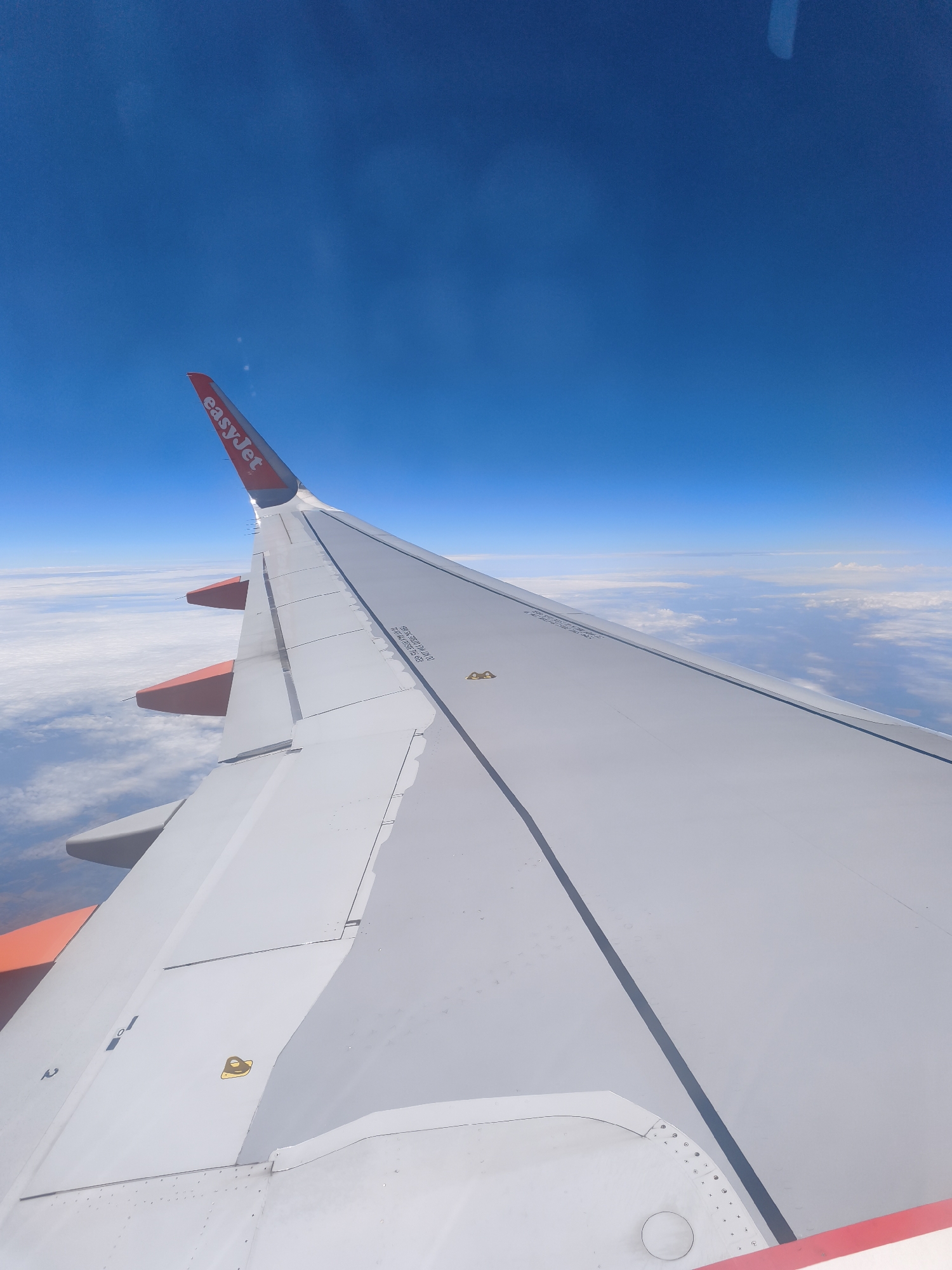 This media file is a photograph taken from the window of an airplane, looking out over a wing. The wing, painted white with orange accents, dominates the foreground and mid-ground. The EasyJet logo is visible in red on the wingtip. The plane is flying above a layer of white clouds, with a clear, bright blue sky above. The sun appears to be high in the sky, suggesting it is daytime. No people are visible. The image conveys a sense of travel and the vastness of the sky. The visible text is "easyJet". The overall impression is one of a clear and sunny flight.