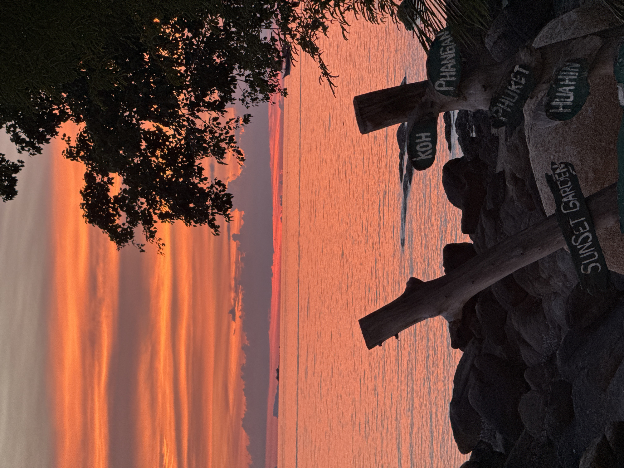 This image depicts a sunset scene in Baan Plai Laem, Thailand. The sky is ablaze with vibrant orange and pink hues, with horizontal bands of clouds. The sea reflects the warm colors of the sky, appearing as a shimmering expanse of orange. In the foreground, dark, rocky terrain leads down to the water. Two weathered wooden posts, one leaning and another more upright, are positioned amongst the rocks. Attached to the upright post are several hand-painted signs indicating various locations: "KOH," "PHANGAN," "PHUKET," "HUAHIN," and "SUNSET GARDEN." A silhouetted tree with dense foliage partially obscures the right side of the frame, framing the sunset. Distant landmasses and a ship are visible on the horizon over the water. The scene is devoid of people.