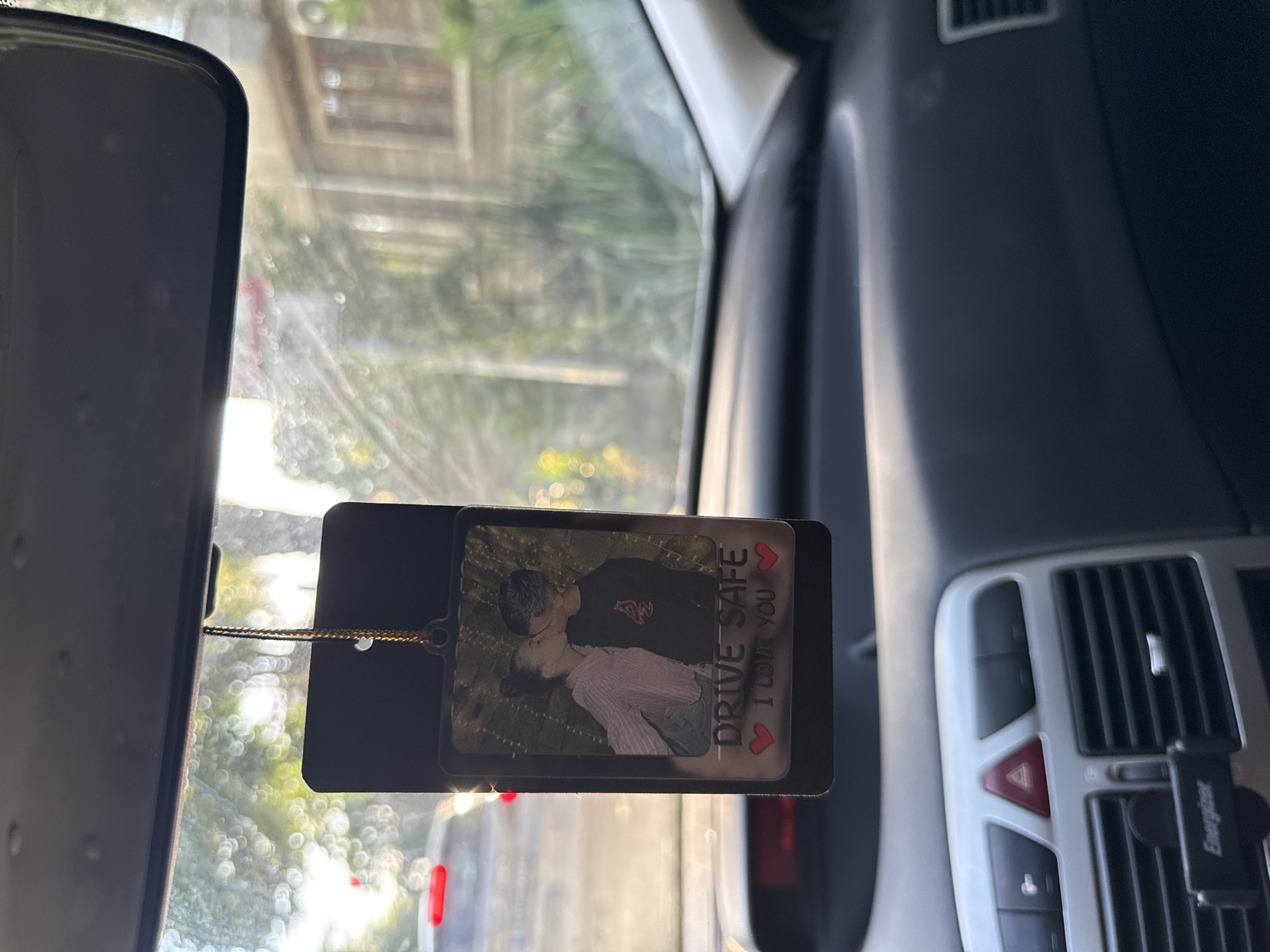 The image shows a close-up of a car's interior, focusing on a personalized air freshener hanging from the rearview mirror. The air freshener features a picture of a young couple kissing, with the text "DRIVE SAFE I LOVE YOU" below. The background, seen through the car's windshield, is blurred but suggests an urban environment with buildings and greenery. Based on the provided information, the location is Stara Zagora, Bulgaria. The car appears to be stopped, possibly in traffic, as indicated by the red taillight of another vehicle visible in the distance. The dashboard of the car is also visible, with the hazard light button illuminated.