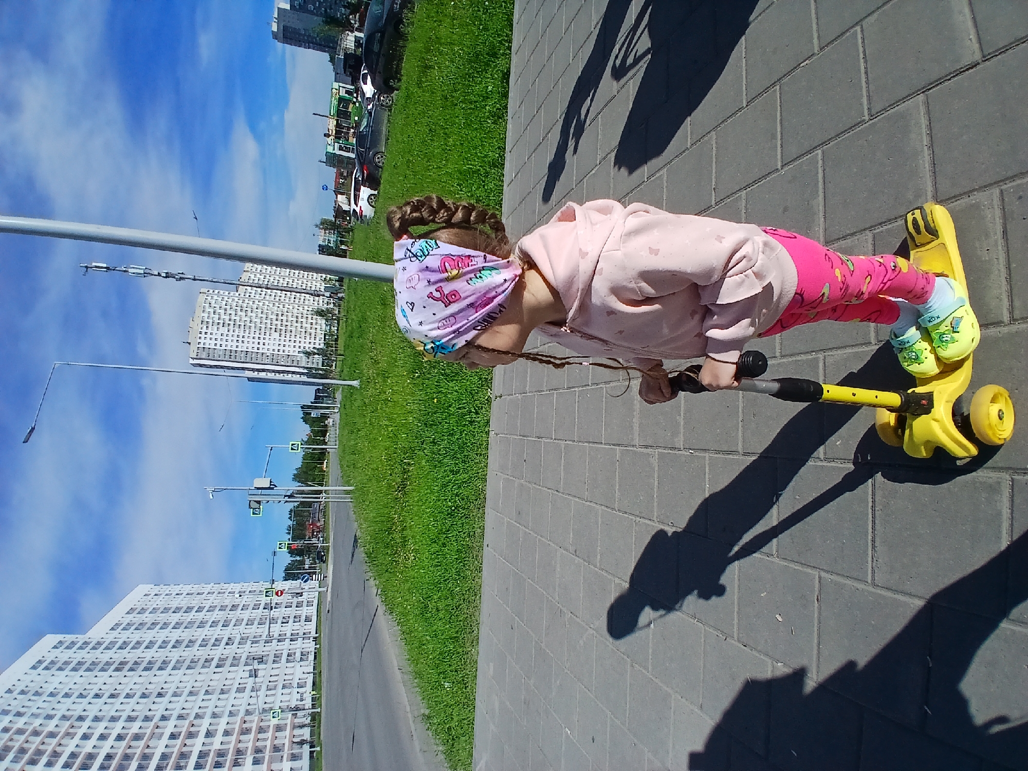 A young girl with braided hair and a bandana stands on a yellow scooter on a paved walkway. She wears a pink hooded sweatshirt and pink leggings with a cartoon print. She is looking to her left, away from the camera. In the background, there are modern apartment buildings, a street with cars, and a grassy area. The sky is blue with some clouds, indicating a sunny day. The scene appears to be in a residential urban area, possibly a newer development in Yekaterinburg, Russia, given the architectural style of the buildings and the presence of pedestrian walkways and streets. The lighting suggests it is daytime.