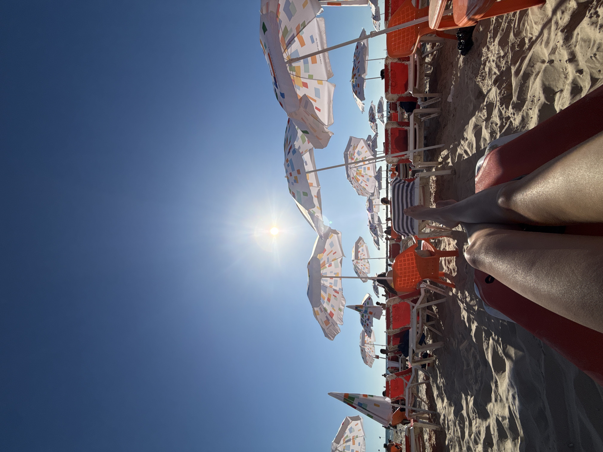 A low-angle, wide shot captures a beach scene in Tel Aviv, Israel, under a clear, bright blue sky. The sun shines intensely, casting lens flare and long shadows across the sandy beach. In the foreground, a person's legs are extended, resting on a red and orange lounge chair, suggesting a relaxed posture. Rows of orange lounge chairs and white beach umbrellas, many with colorful geometric patterns, are set up on the sand. In the mid-ground, several people are visible, some sitting in chairs or walking along the beach. The background shows the open expanse of the sky and a hint of the sea. The overall atmosphere is one of a sunny day at a popular beach.