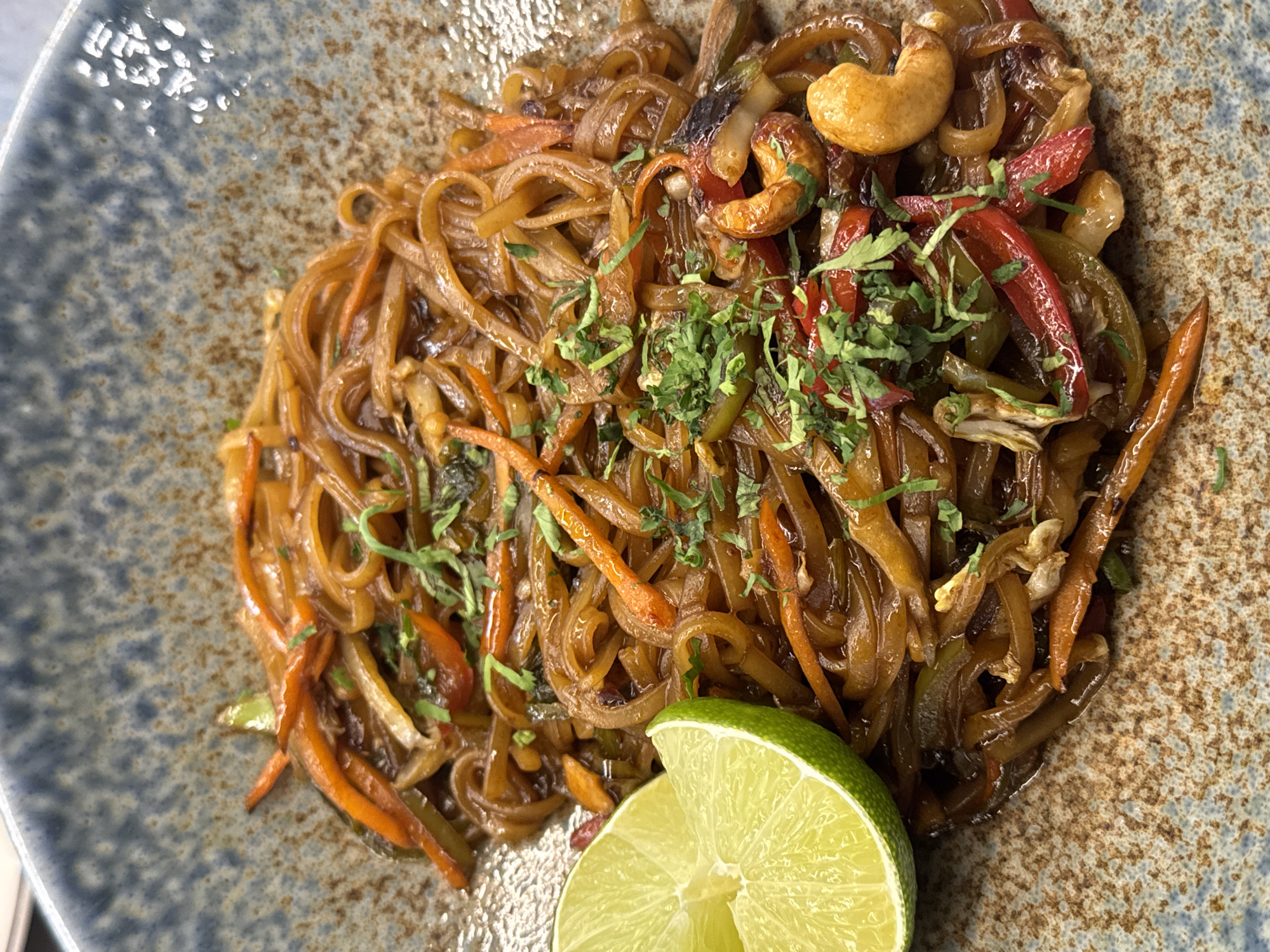 A close-up, overhead shot shows a serving of stir-fried noodles in a speckled, blue-rimmed ceramic bowl. The noodles are thin and coated in a dark, glossy sauce, interspersed with strips of orange carrot, green and red bell pepper, and whole cashews. Chopped green herbs are sprinkled over the dish. A lime wedge rests on the left side of the bowl. The background is out of focus, suggesting an indoor setting. The image does not contain any recognizable landmarks or details indicating it is in Sofia, Bulgaria.
