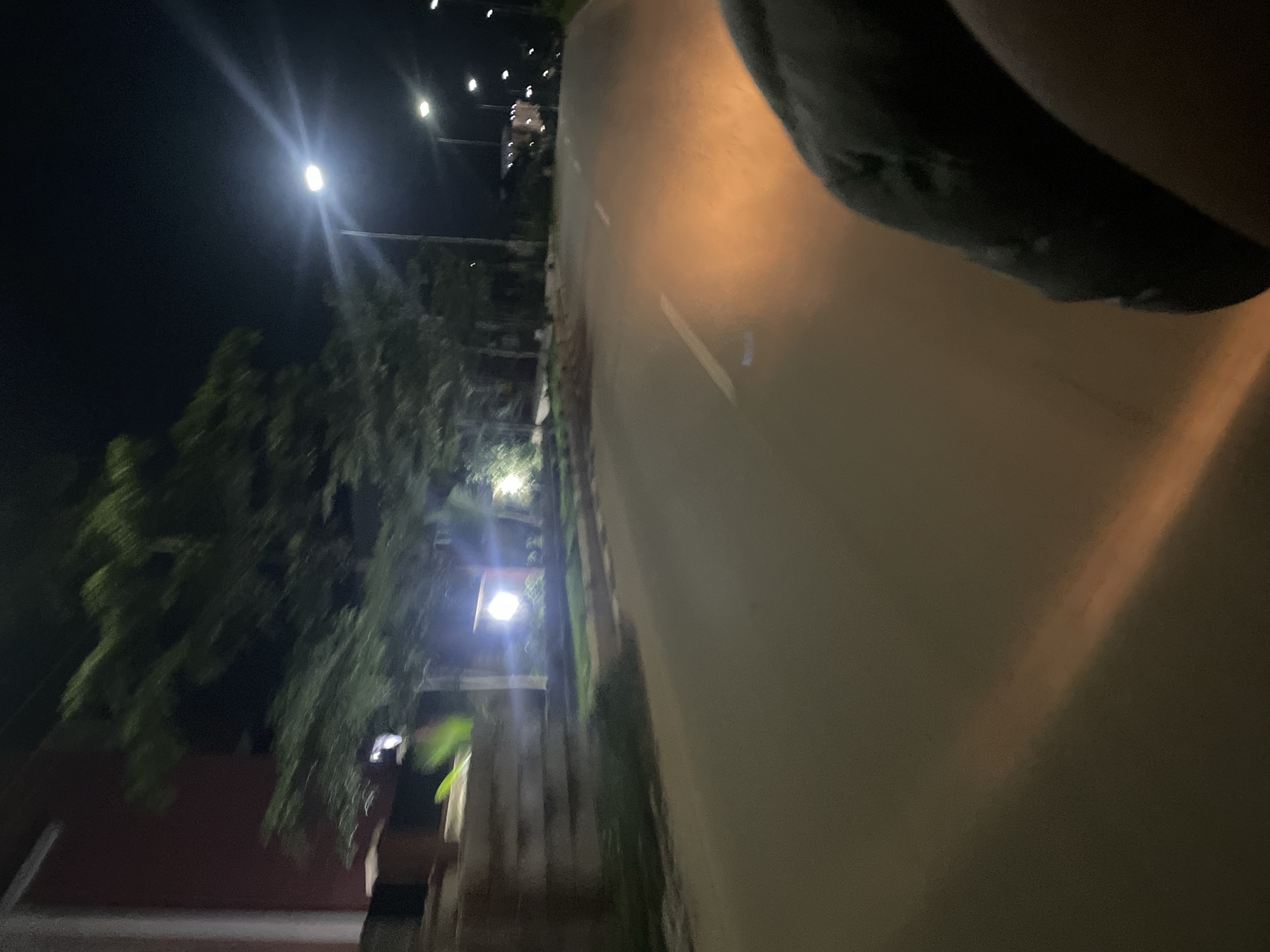 The image captures a nighttime view from the perspective of someone on a moving vehicle on a paved road in Guzape, Nigeria. The scene is illuminated by streetlights and the headlights of an approaching vehicle, casting a warm orange glow on the asphalt. Trees with dense foliage line the side of the road, with a building visible in the background. The vehicle's occupant is partially visible in the foreground, their dark clothing suggesting they are wearing a jacket or coat. The road has painted white lines marking the lanes. The overall atmosphere is dimly lit, characteristic of a nocturnal urban or suburban setting.