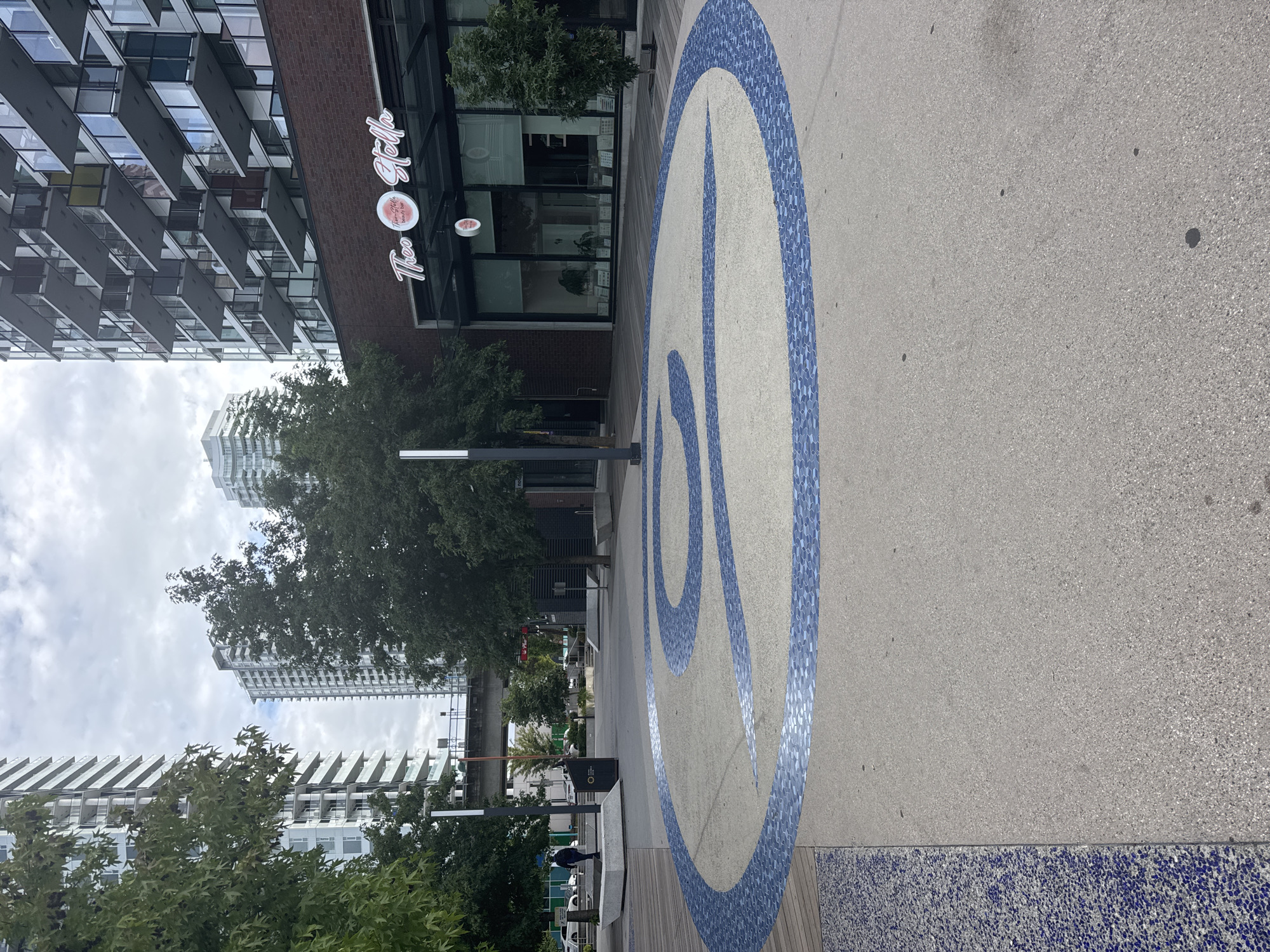 The image depicts an outdoor public space with a distinctive circular mosaic design on the ground, featuring a blue, swirling pattern. This mosaic is set within a concrete plaza, bordered by wooden decking on the left. In the background, modern high-rise buildings are visible, suggesting an urban setting. A brick building with a commercial storefront, identified by a sign reading "Theo + Stella beauty bar," is also prominent. Trees are planted throughout the area, adding greenery to the scene. A person is walking in the distance on the left side of the image. The sky is overcast, indicating a cloudy day. The overall impression is a contemporary urban plaza with artistic elements. This image is likely taken in Surrey, Canada, based on the architecture and likely context.