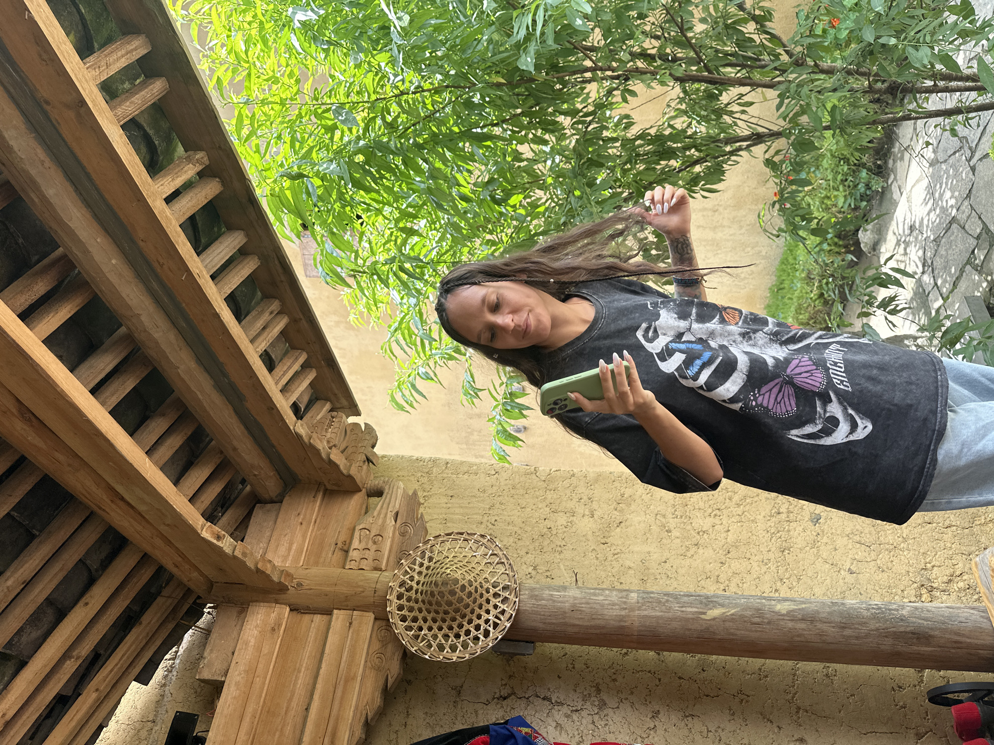 A woman with long brown hair partially braided is standing outside, partially framed by traditional wooden architecture and green foliage. She is holding a phone and looking down at it. She is wearing a dark t-shirt with a graphic print and light-colored jeans. A woven conical hat is mounted on a wooden beam. The setting appears to be in the Lũng Cú Commune in Vietnam, implied from traditional architecture.