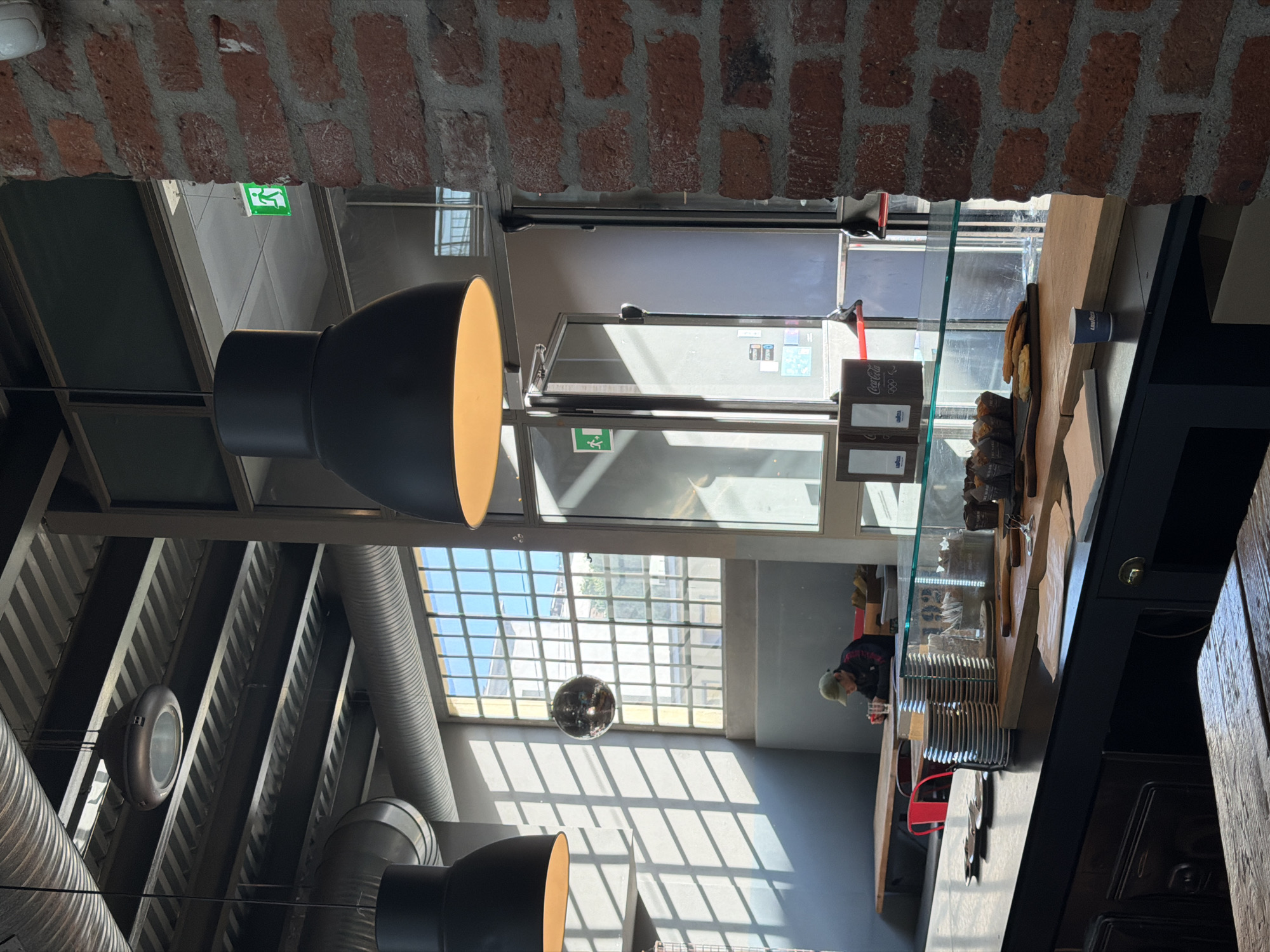 View looking into a cafe or food establishment in Unknown, Unknown, featuring industrial-style design elements. The foreground is framed by a weathered brick archway. Two large, black pendant lamps with warm-toned interiors hang from the ceiling. Exposed ductwork and support beams add to the industrial aesthetic. A disco ball is suspended from the ceiling. Natural light streams in through a window with a grid pattern. The service area features a display case with food items, a service counter, and a person visible behind the counter.