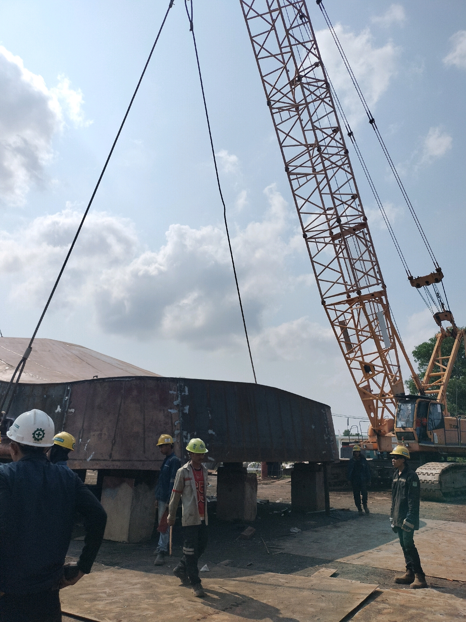 In Batam, Indonesia, a large cylindrical metal structure, likely part of a tank or vessel, is being lifted by a massive yellow crane. The structure is suspended by thick cables. Several construction workers, all wearing hard hats, are visible on the ground, some observing the lift and others working. The environment appears to be an industrial or construction site, with dirt and metal plating on the ground. The sky is partly cloudy, and trees are visible in the background.