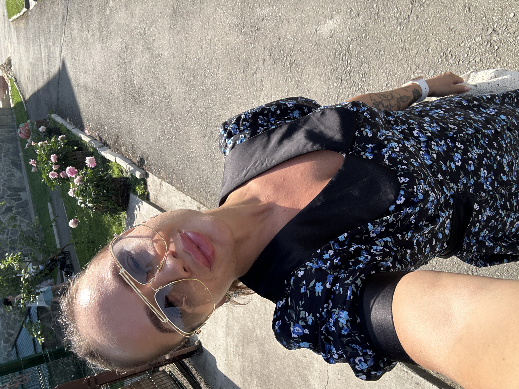 This is a portrait-style, slightly tilted photograph of a person outdoors. The subject, a person with light skin and blonde hair, is wearing large, gold-rimmed sunglasses and a dark floral-print dress with blue flowers. They are also wearing a white smartwatch on their left wrist. A tattoo of a floral design is visible on their left forearm.
The setting appears to be a paved area, possibly a pathway or driveway, with a shadow cast on it from an unseen object, suggesting it is daytime. To the left of the paved area, there are some potted plants with pink flowers and greenery. Further in the background, there's a stone wall and more vegetation. The lighting suggests it is a sunny day, possibly late afternoon or early morning due to the long shadow. The overall mood seems casual and relaxed. There is no visible text in the image.