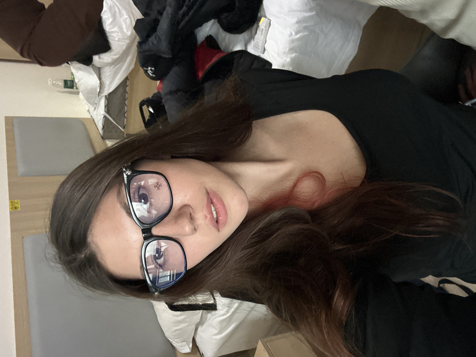 An image depicts a single female individual in a mid-shot, facing directly towards the camera, situated within an interior room identified as a hotel room in Huangdao District, China. The subject has long, dark brown hair, with a distinct reddish or pink streak visible on the left side of her hair (viewer's right). She is wearing black-framed glasses with decorative silver-toned elements on the temples; a red cross symbol is visible as a reflection on the left lens. She is wearing a black, scoop-neck garment.
The background shows a disheveled room environment. To the subject's left, a portion of a bed with white linens and a white pillow is visible, adjacent to a light-colored wooden bedside table. The headboard behind this bed features light wood paneling with a grey padded section, above which a small yellow rectangular sign is affixed. To the subject's right, another bed or surface is heavily cluttered with white linens, several dark-colored articles of clothing (including what appears to be a black jacket), a clear plastic water bottle, and a white remote control. A large brown fabric object is also present in this area. The overall scene suggests a lived-in and somewhat untidy domestic or temporary lodging space.